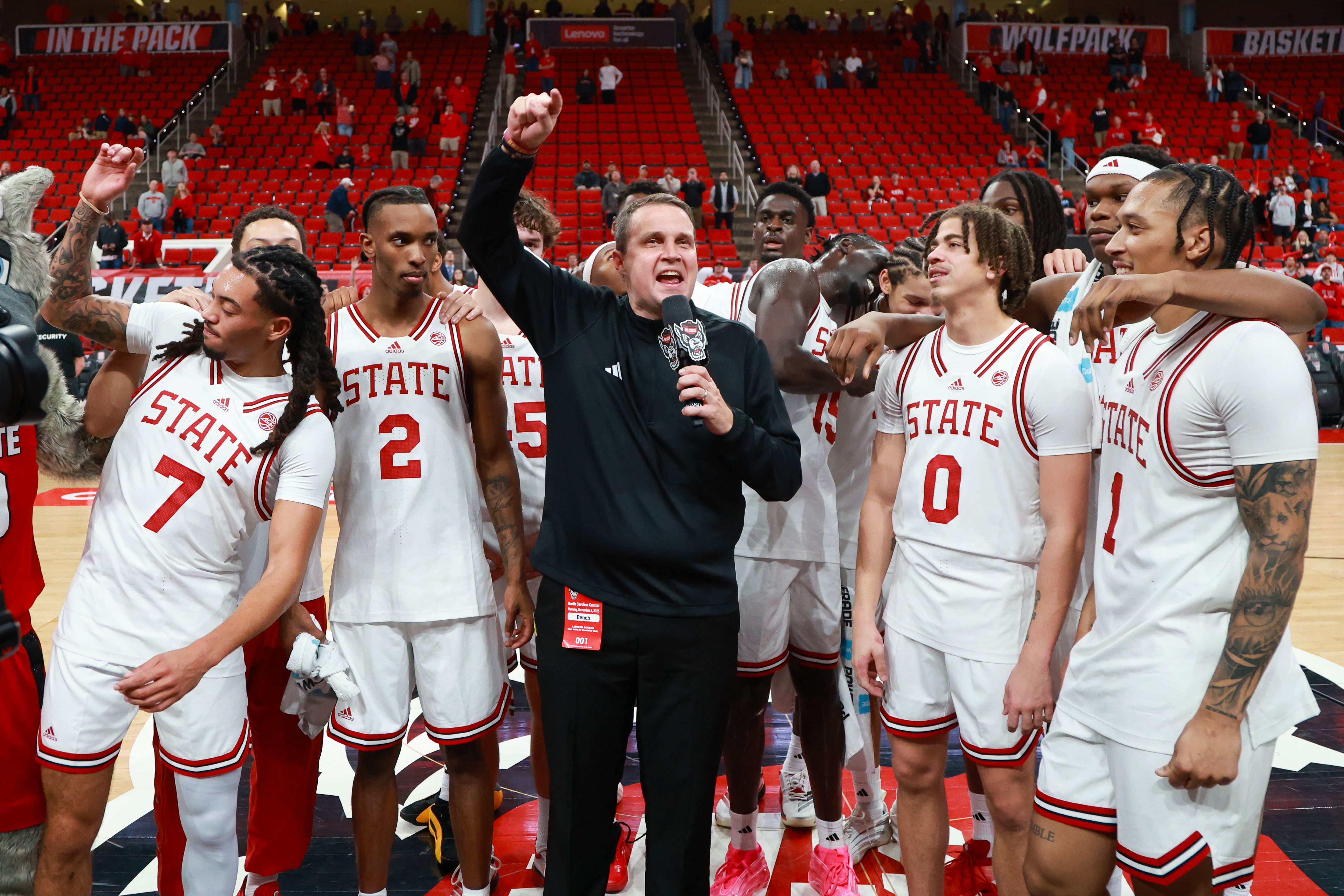 Basketball team in white "STATE" jerseys celebrating on court, coach in black holding microphone with raised fist, red stadium seats and signs "IN THE PACK", "WOLFPACK" in background.RALEIGH, NC - NOVEMBER 03: North Carolina State head coach Will Wade speaks to the crowd after the college basketball