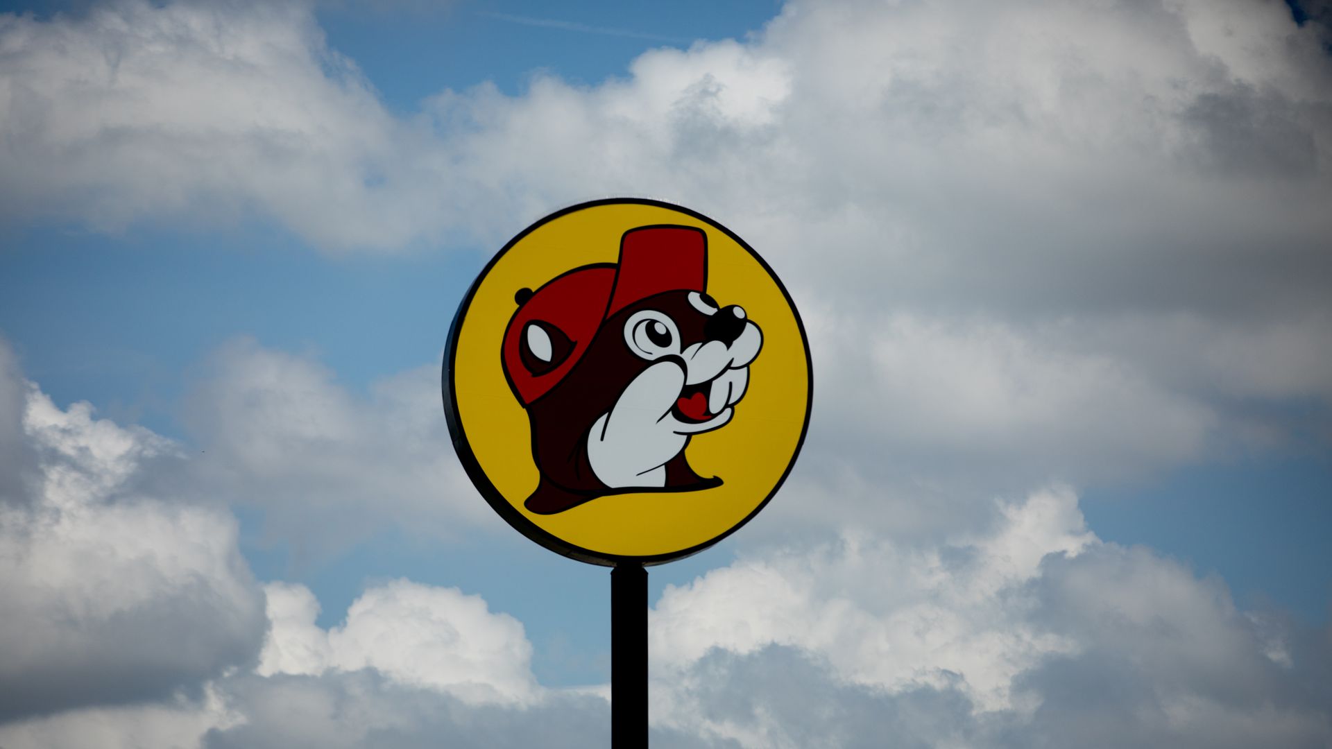 Photo of a highway sign with a cartoon beaver with a red hat. Clouds and blue sky in the background.