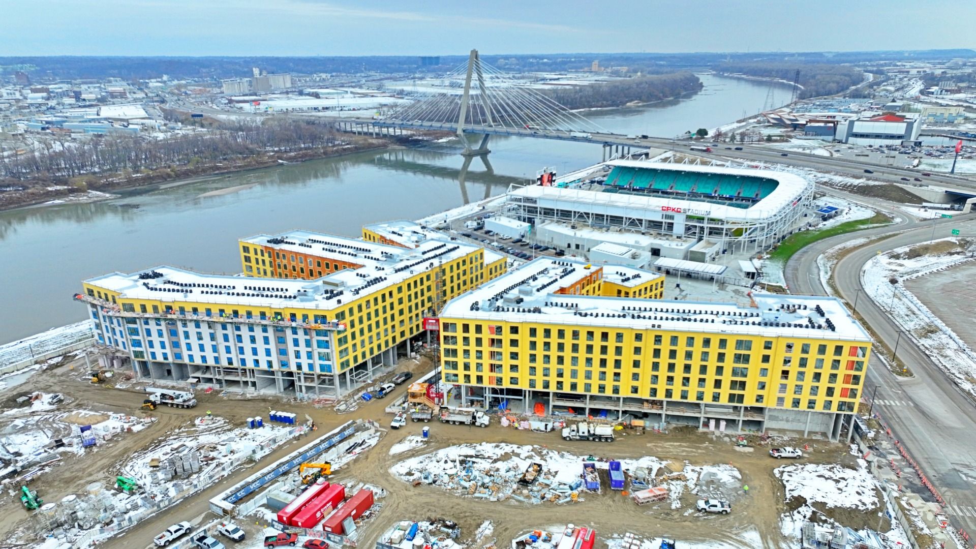 Aerial view of large yellow and blue buildings under construction by a river, with a cable-stayed bridge and CPKC Stadium in the background, on a partly snowy day.