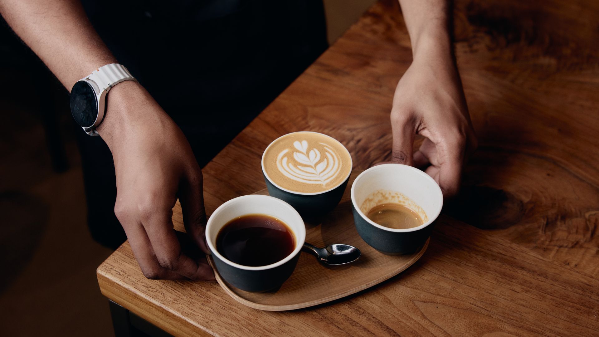 Two hands hover over a wooden table with a circular tray of coffee cups. A latte with latte art rosette sits in a dark cup; one wrist shows a white watch, others in dark sleeves.