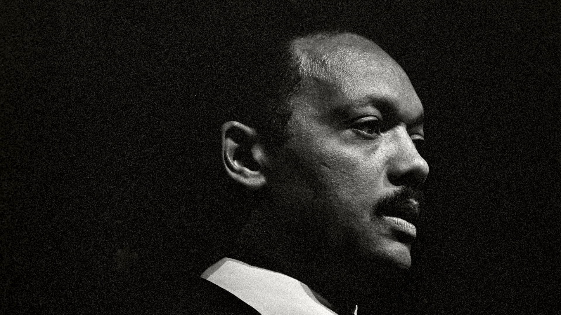 Black and white close-up portrait of a man in a suit looking right, with a serious expression, against a dark background.