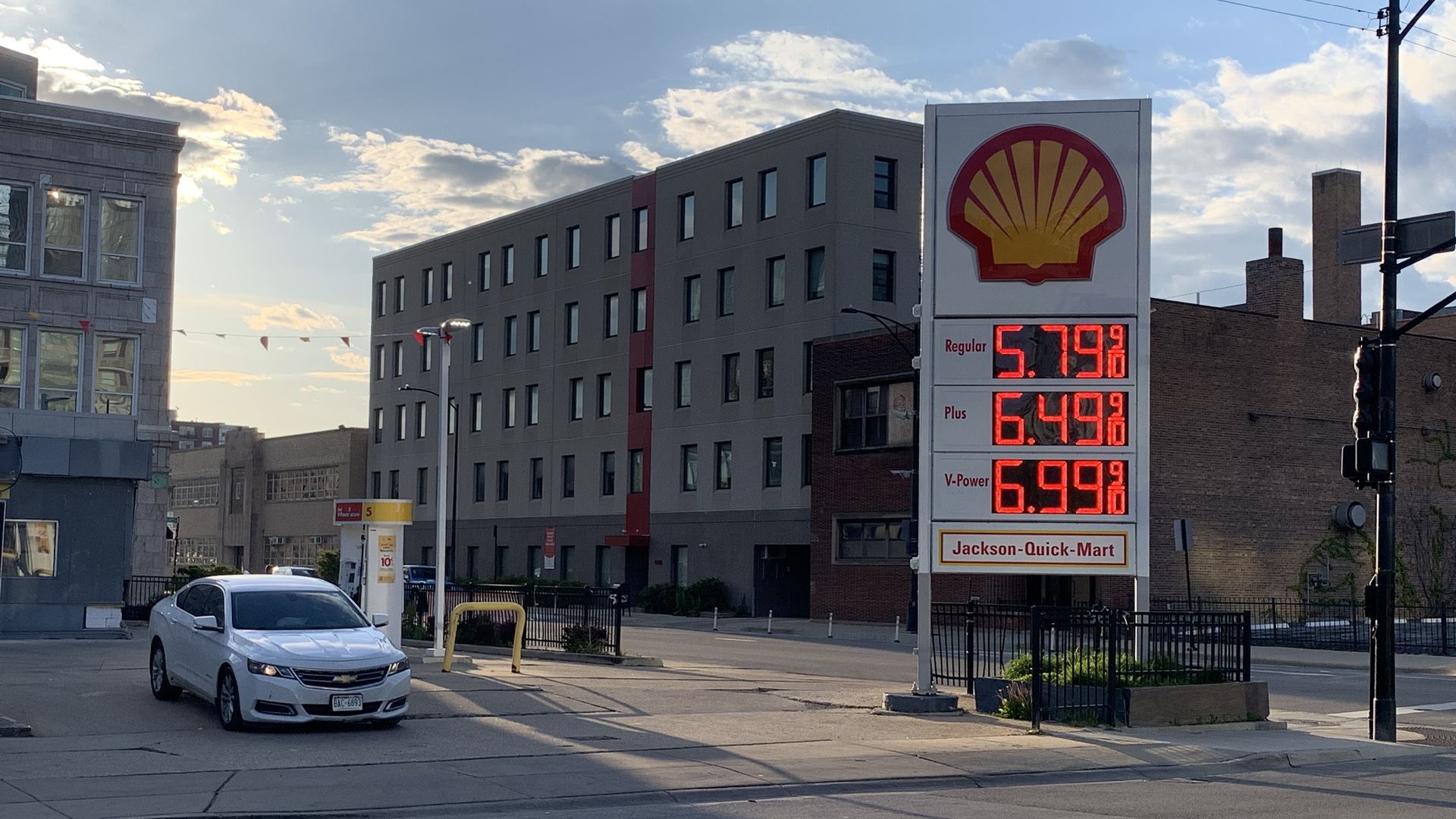 Shell gas sign with red digital prices dominates right side; white car in foreground; gray modern apartment buildings; blue sky with scattered clouds.