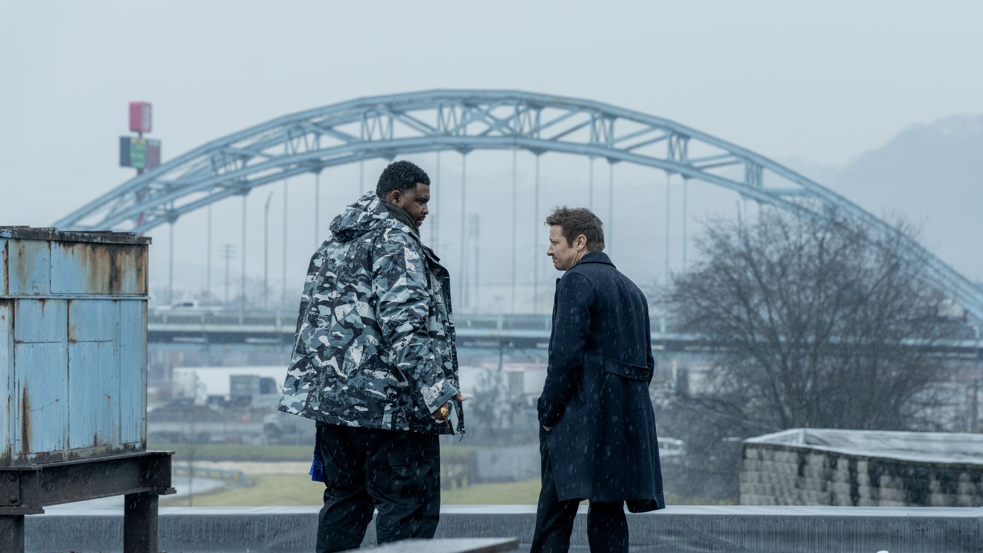 Tobi Bamtefa looks down at Jeremy Renner while they appear to be talking outside in the cold.