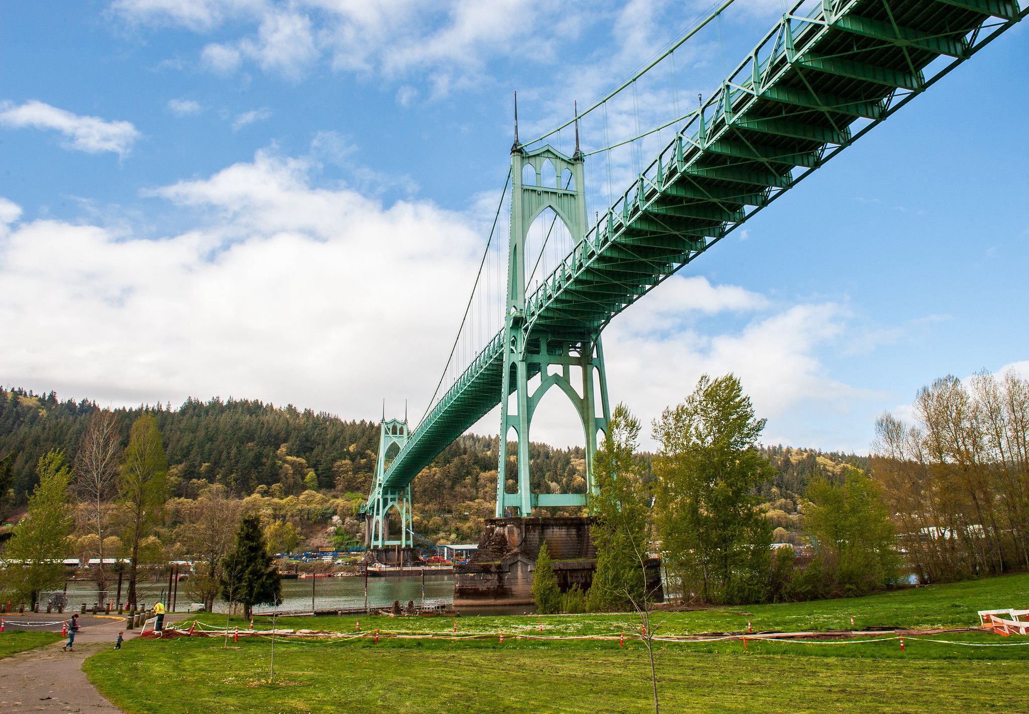 Green metal suspension bridge spans a river under a bright blue sky with clouds; a grassy park and tree line fill the foreground, with a few people walking near the water.