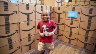 A boy in a red shirt stands in front of stacked Amazon boxes