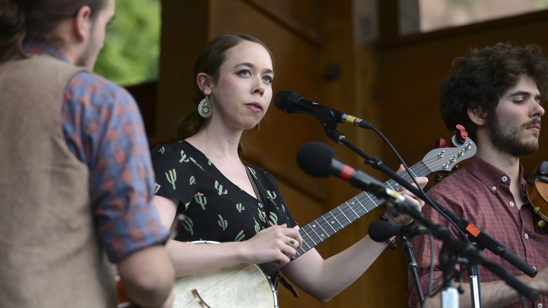 Female singer in a cactus-pattern dress and green earrings plays an acoustic guitar at a microphone on an outdoor wooden stage, flanked by two male musicians.