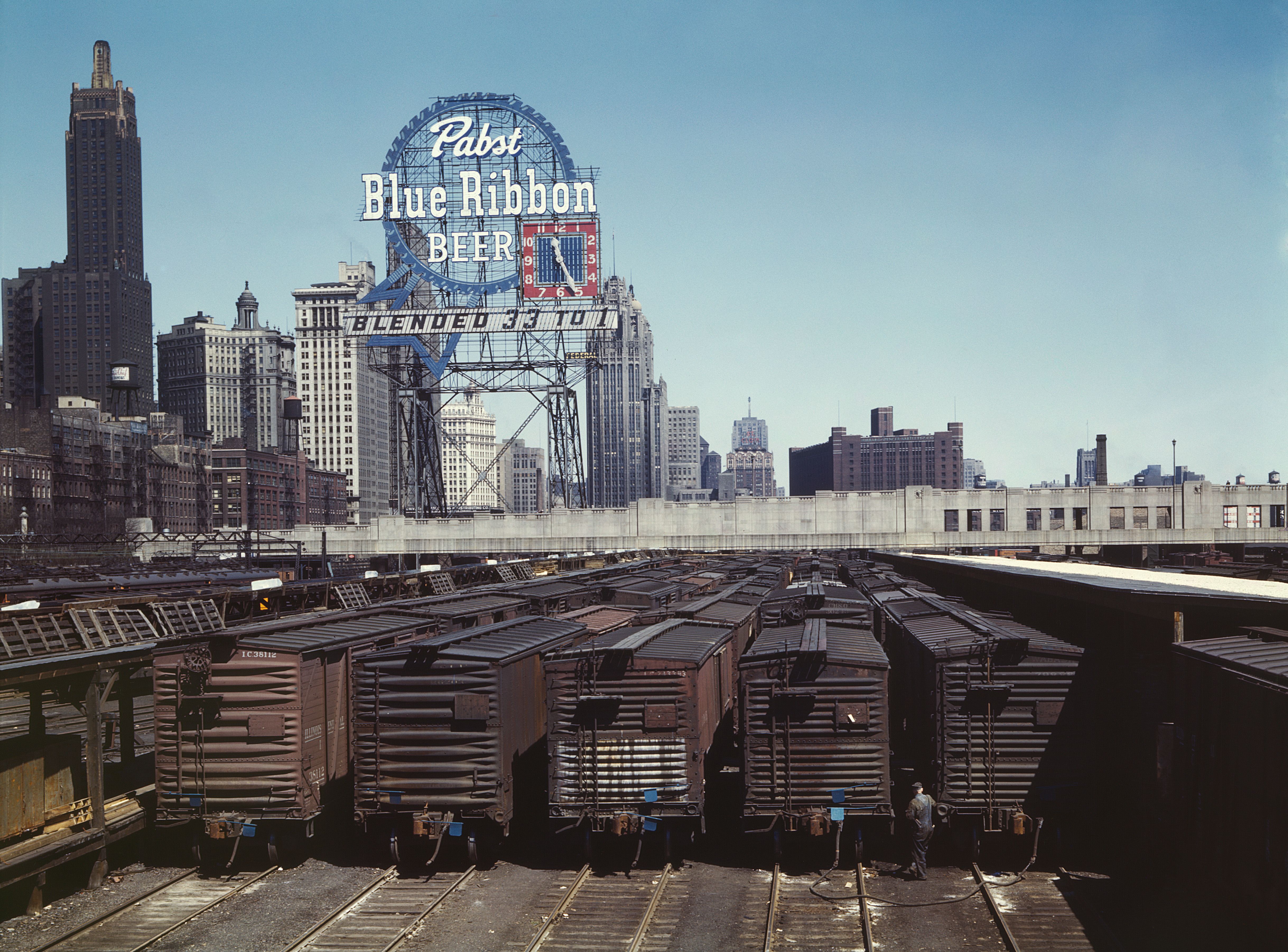 Urban skyline with a large Pabst Blue Ribbon Beer sign on a steel framework above a building, tall towers nearby, and a rail yard with stacked freight cars in the foreground under a blue sky.