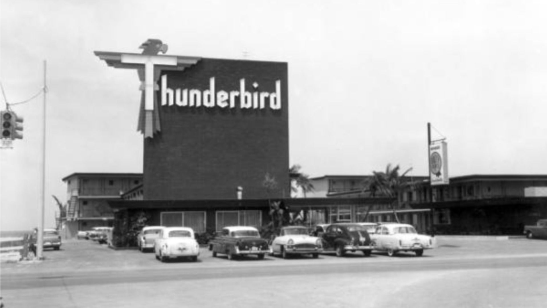 A black-and-white image of a hotel on the beach with a large sign that says "Thunderbird." The T is decorated to look like a bird.
