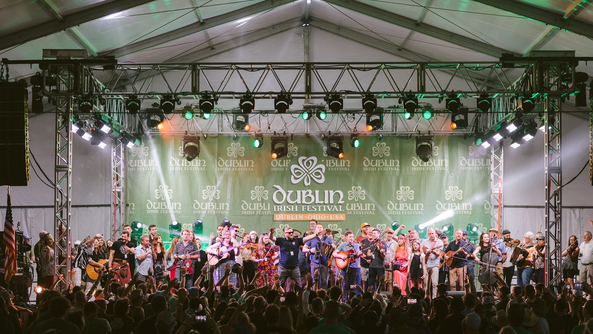 Wide stage at a Dublin Irish Festival show: many musicians play guitars, fiddles, and accordions under green lights, while a large audience watches from the foreground.