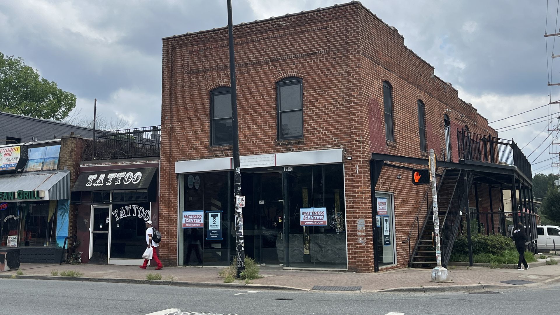 Brick corner building with large storefront windows housing a tattoo shop and a mattress center sign. A tall street lamp in the foreground, overcast sky, and a pedestrian in red pants walking by.