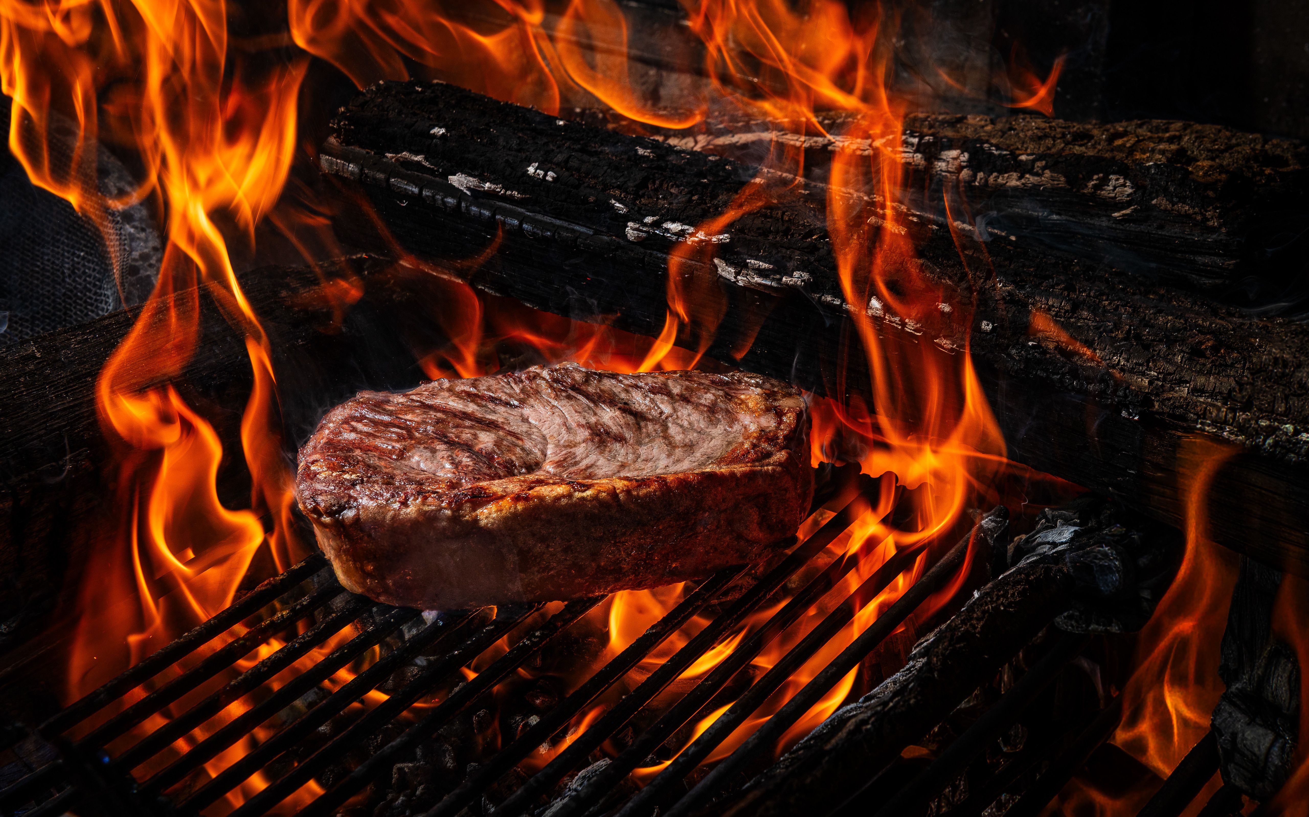 A thick, seared steak rests on a hot grill as bright orange flames lick the sides, with dark charcoal and charred logs visible in the background.