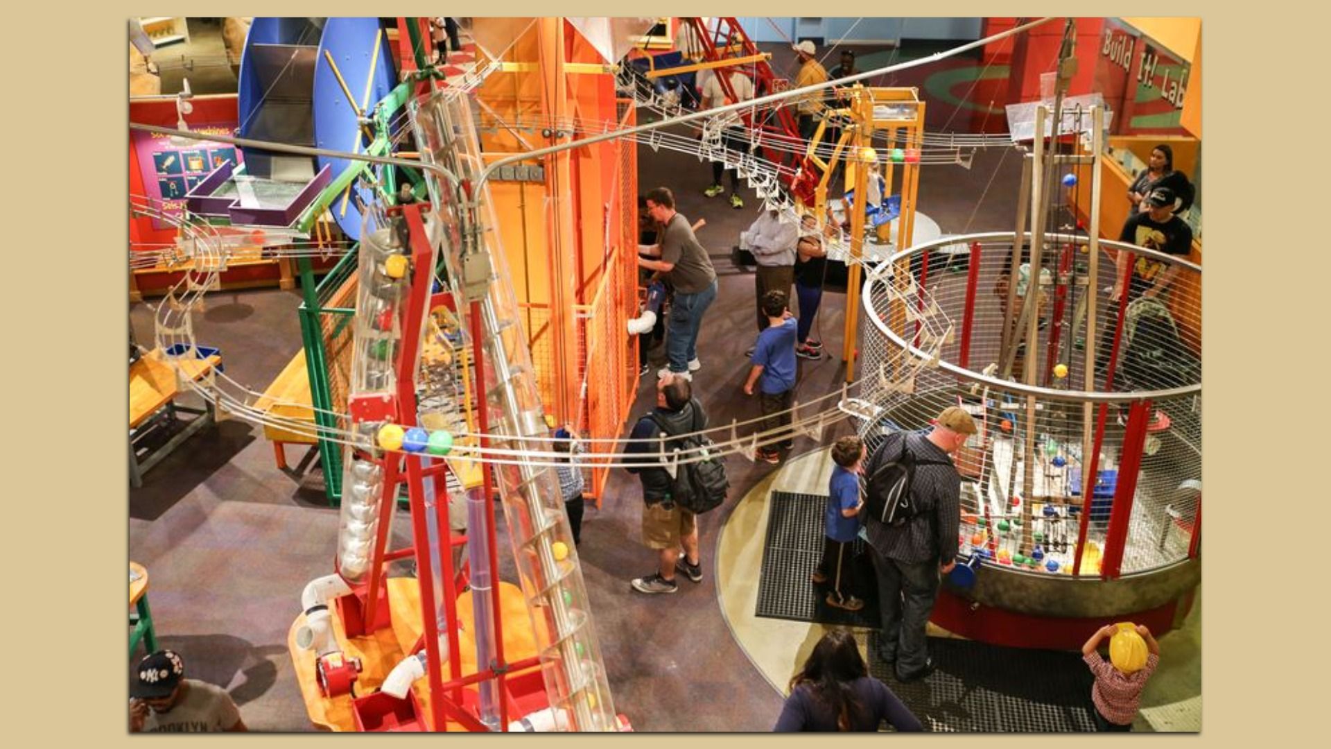 Colorful indoor science exhibit with tangled metal tracks, cables, and orange supports. People of various ages move around a multi-level, spiraling kinetic structure.