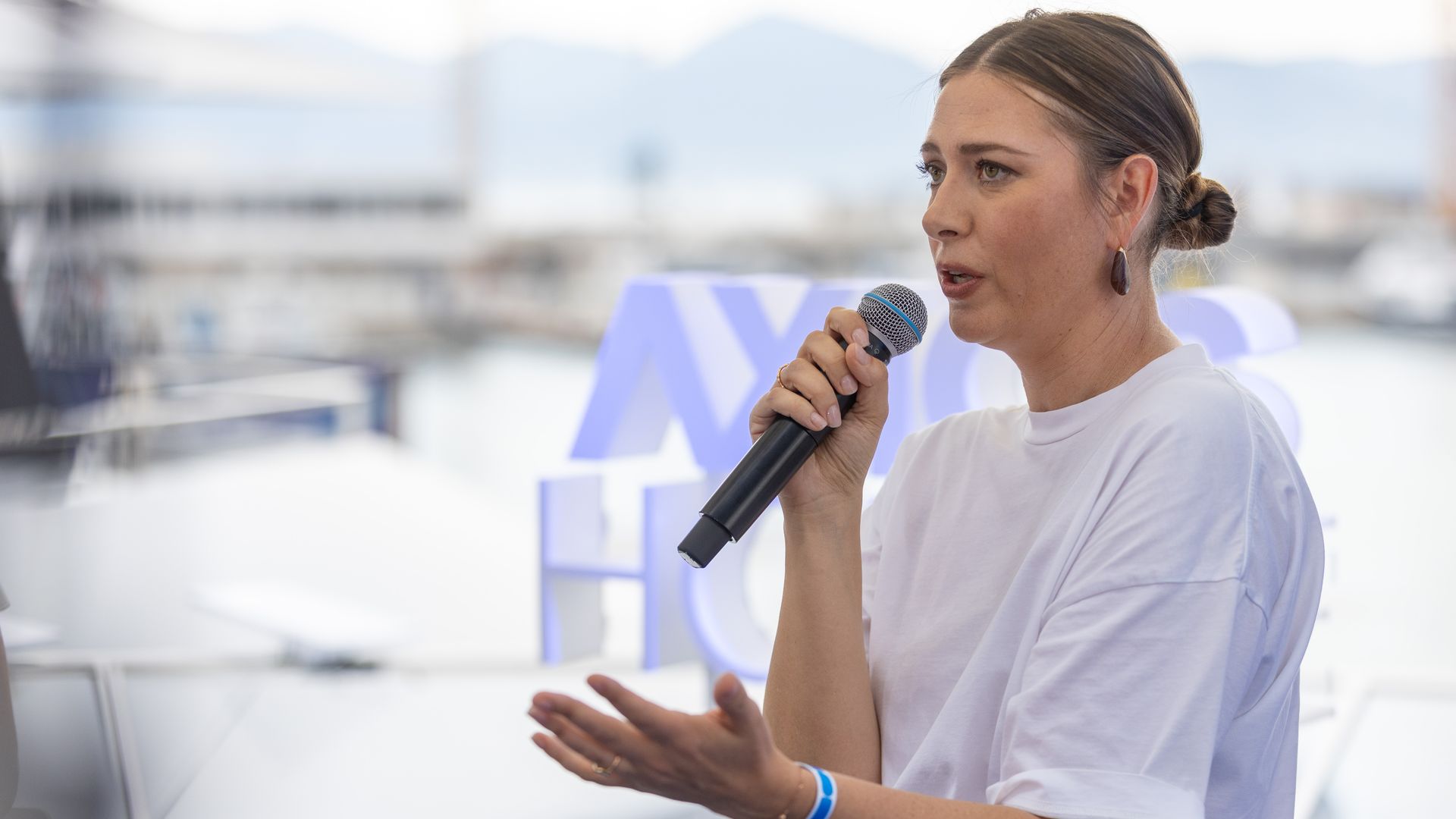 A woman in a white shirt speaks into a handheld microphone at a bright event, her brown hair in a bun and wearing a purple teardrop earring, with a blurred blue background.