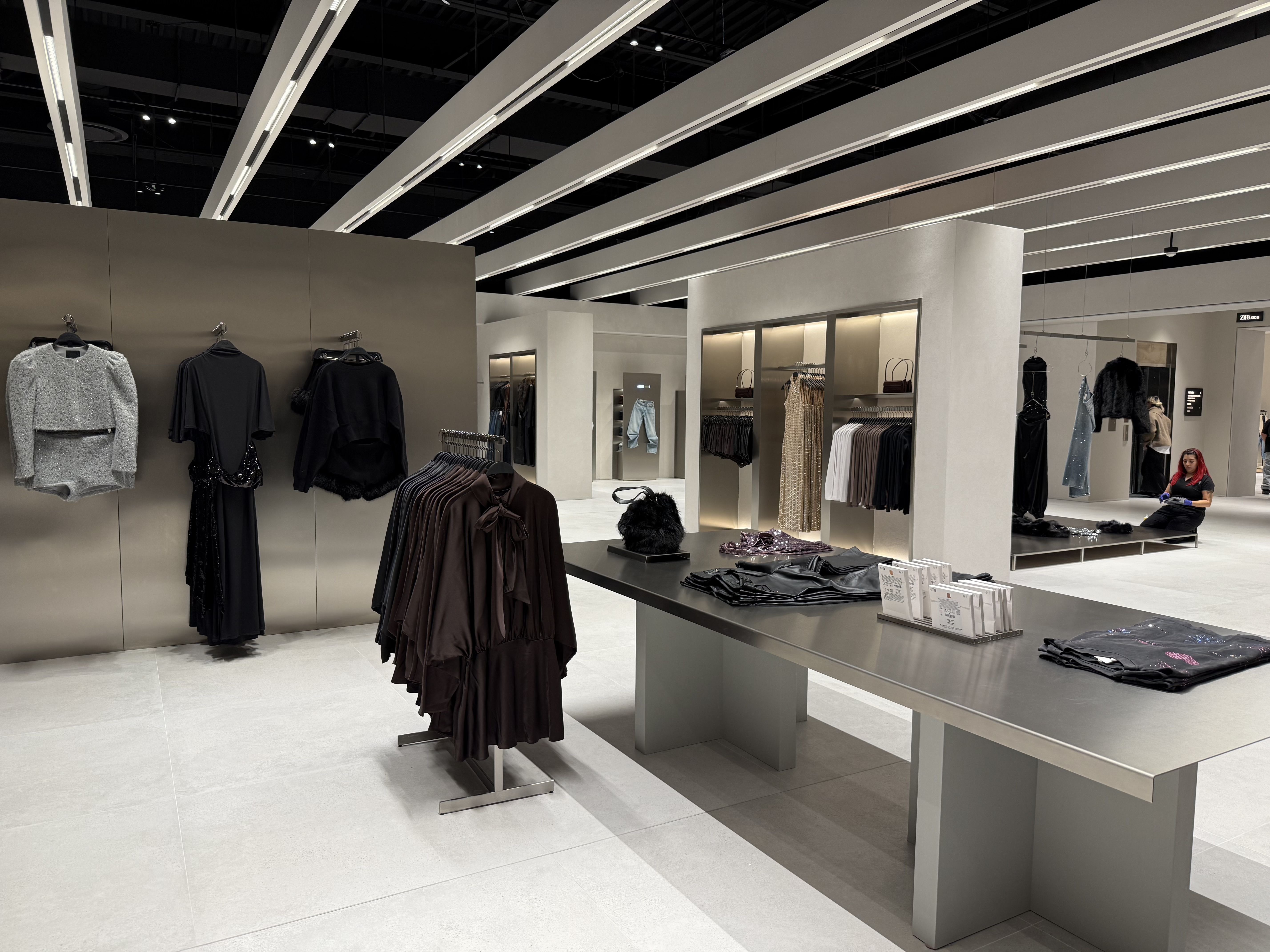 Interior of a modern clothing store with black and neutral-toned garments on racks and tables, light-colored floor, and a woman with red hair sitting on the floor near a large mirror.