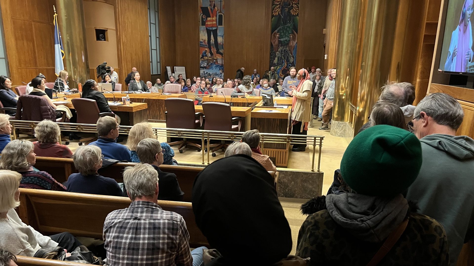 A long line to reach a podium at a city council meeting in a wood-paneled chamber with a large audience