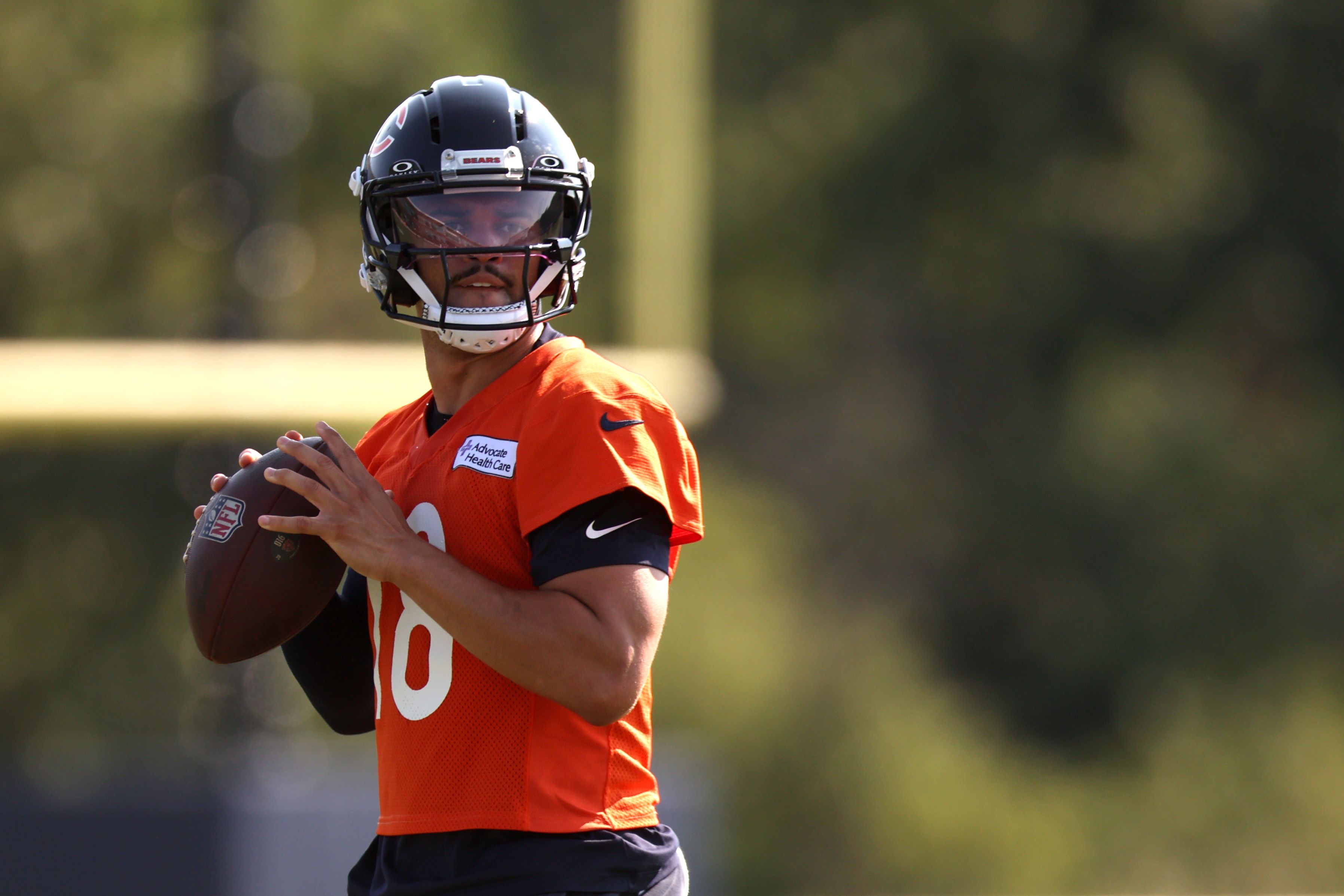 Quarterback in an orange jersey and black helmet with visor, holding a football and looking ahead on a blurred field.