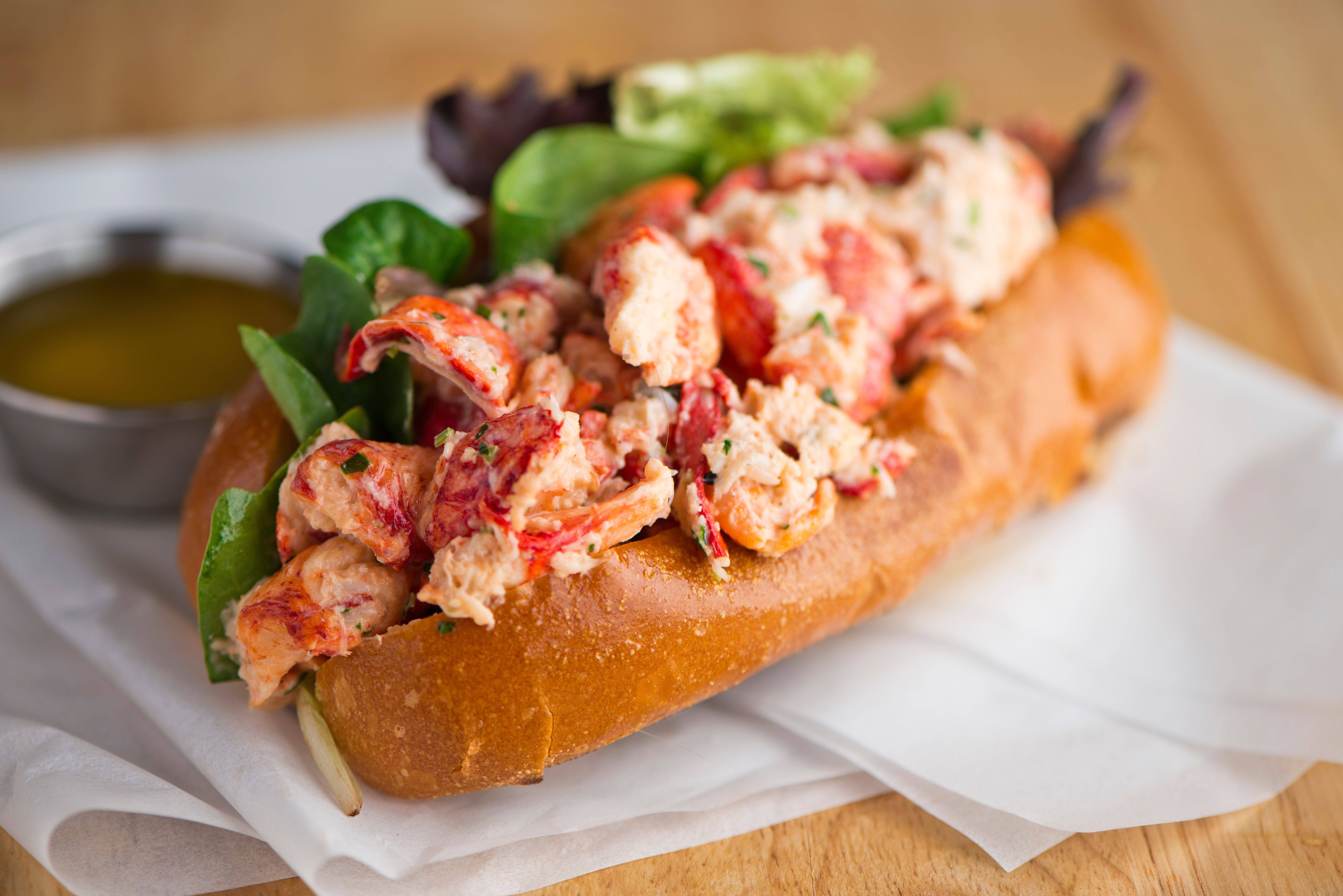 Close-up of a lobster roll on a toasted bun, stuffed with pink lobster meat and herbs, with leafy greens and a small dipping sauce cup in the blurred background.