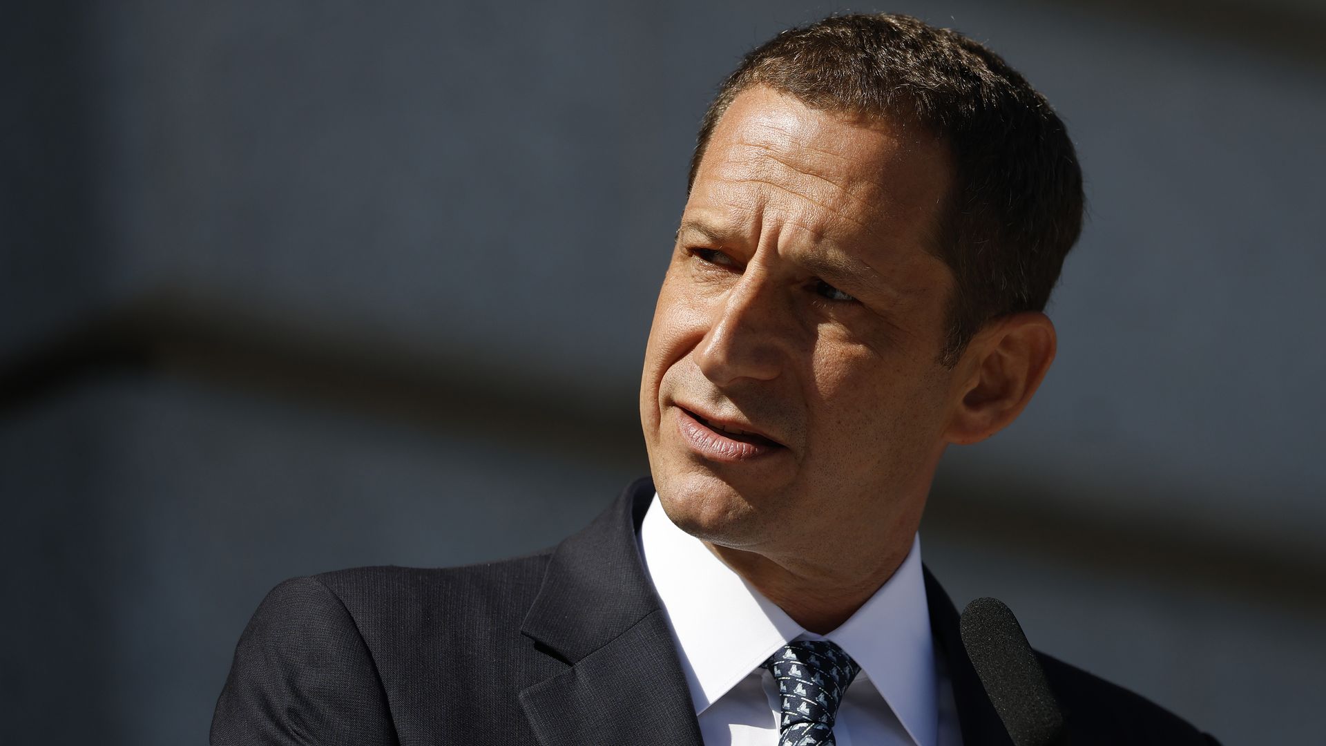 Man in dark suit, white shirt, and patterned tie speaking into microphone, looking to the side against a blurred gray background with sunlight casting shadows on his face.
