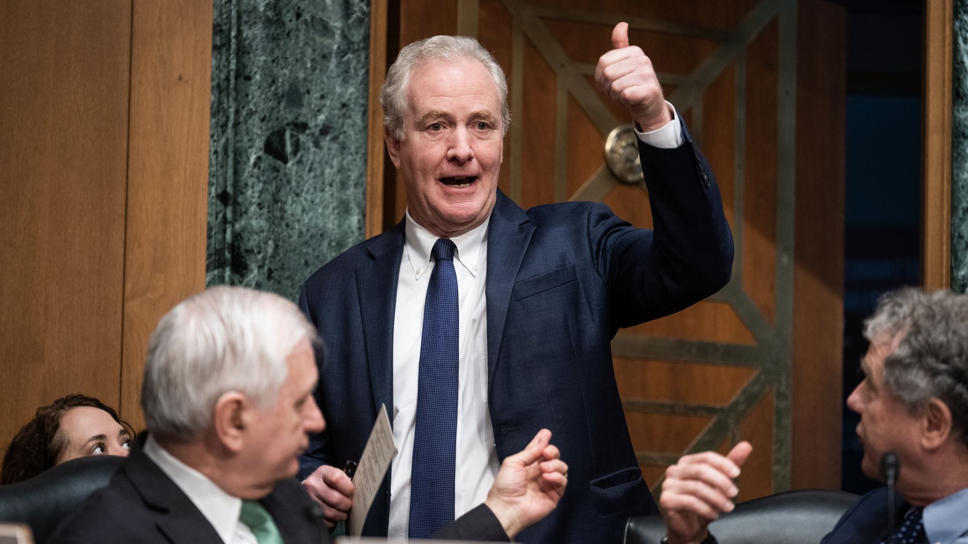 Sen. Chris Van Hollen, wearing a dark blue suit jacket, white shirt and blue tie, giving a thumbs up in a Senate committee chamber.