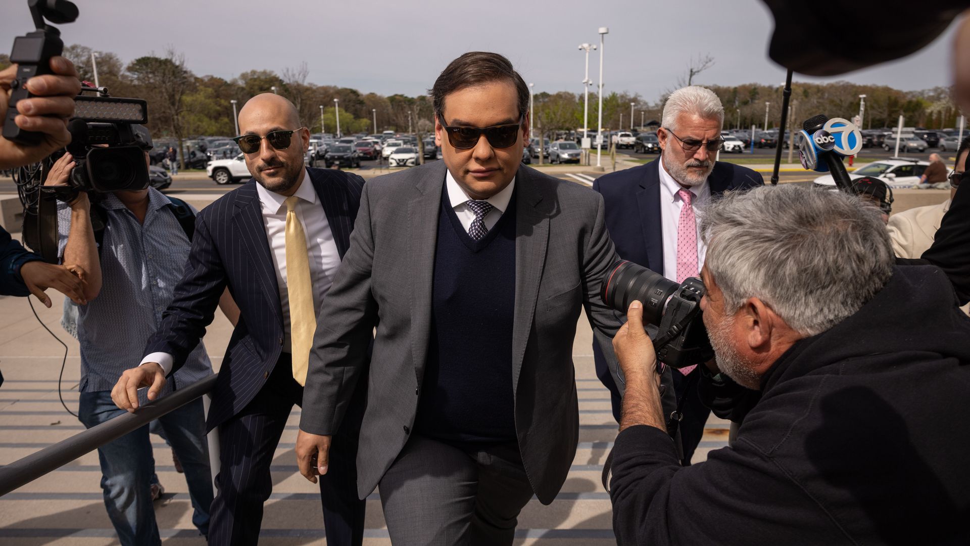 A person in a gray suit and dark sweater, wearing sunglasses, walks amid a crowd of reporters and photographers; cameras and microphones surround him in a parking lot.
