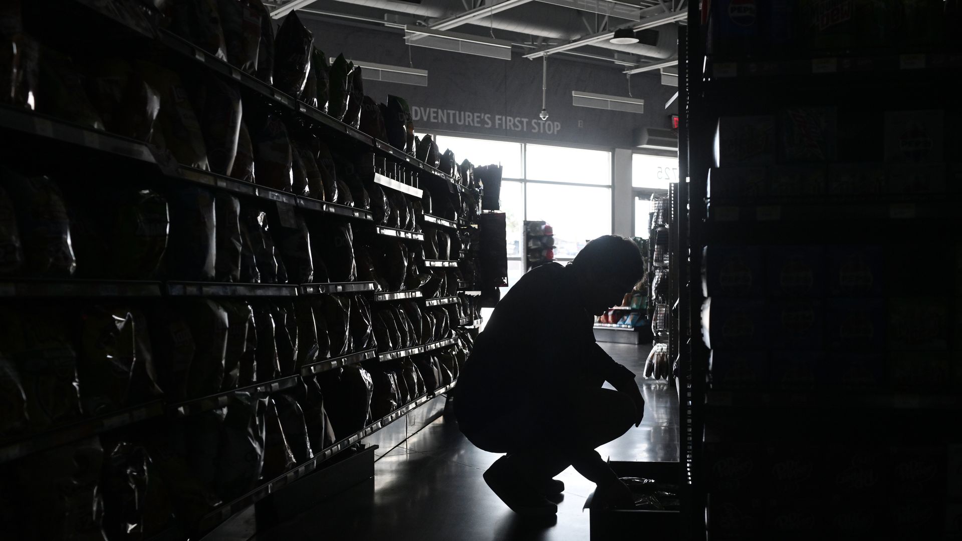 Silhouette of a person crouching between store shelves stocked with snacks and drinks, with bright daylight coming through large front windows in the background.