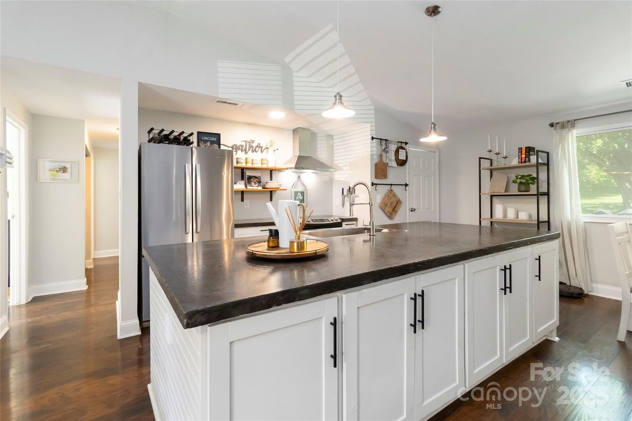 Modern kitchen with white cabinetry and a large dark countertop island, stainless steel fridge, open shelves, wood floor, and natural light from a window with sheer curtains.