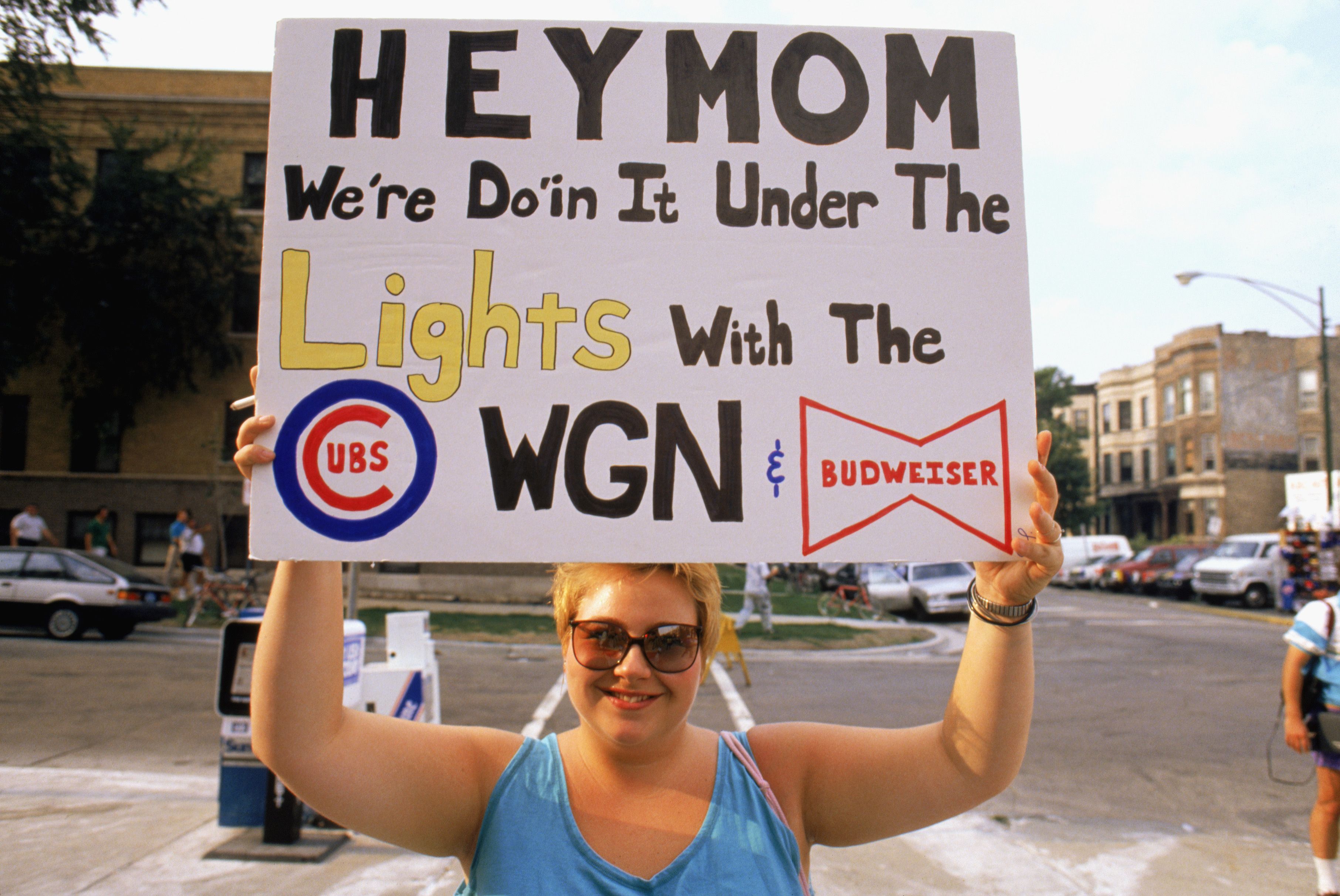A person wearing sunglasses and a blue sleeveless top holds a sign saying, "HEY MOM We're Doin It Under The Lights with the Cubs, WGN & Budweiser" in a street with cars and buildings in background.