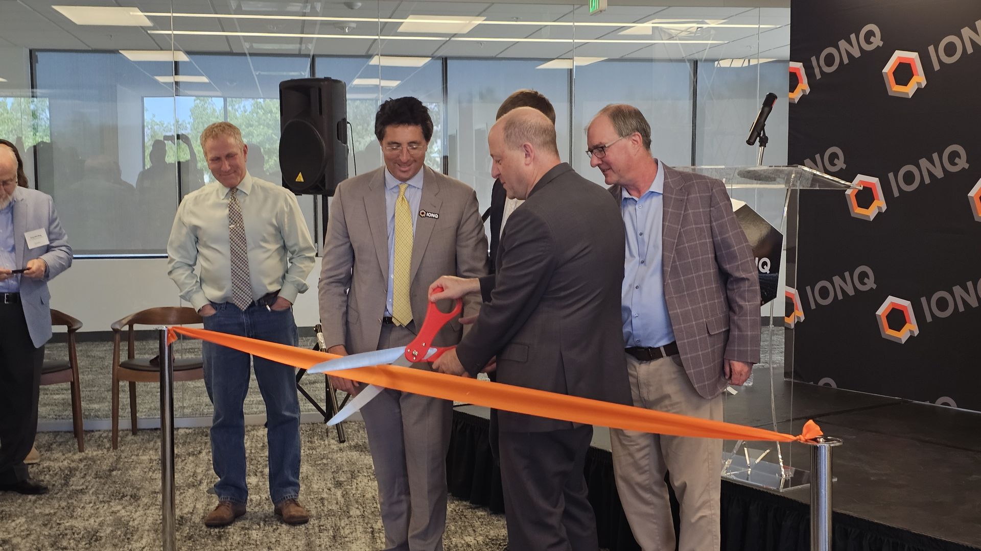 Group of suited professionals at a ribbon-cutting ceremony cutting an orange ribbon with oversized scissors. A black IONQ backdrop with hex logos and a clear podium.