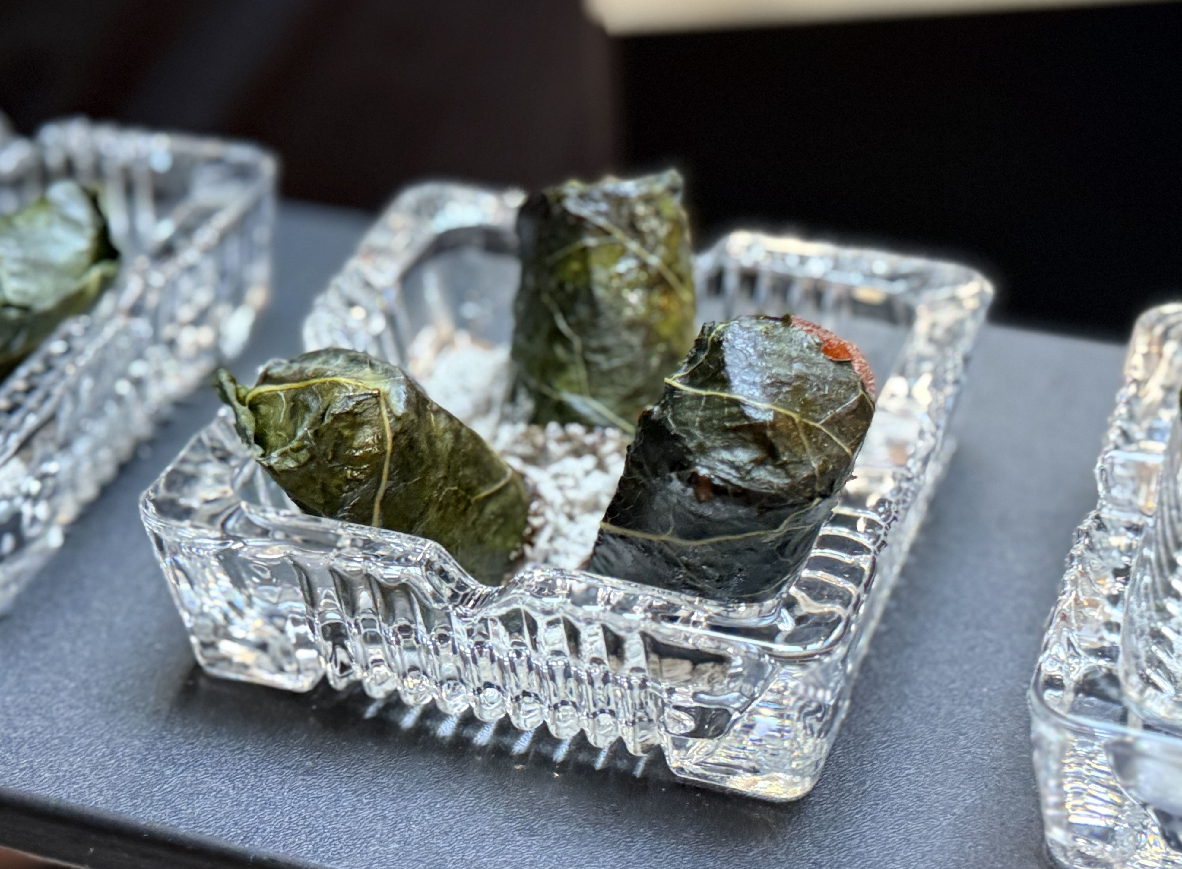 Close-up of dolmas (stuffed grape leaves) arranged in clear ice trays on a dark surface; green leaves with strings and salt granules around them.