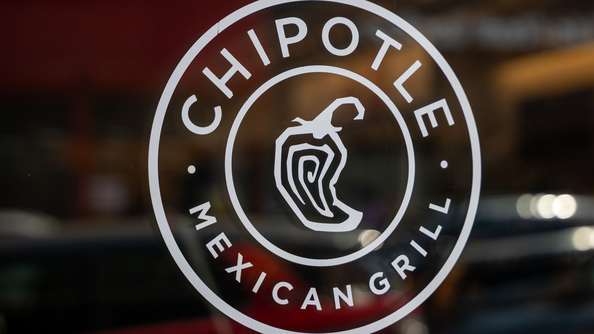 Close-up of a white Chipotle Mexican Grill logo on glass: a circular emblem with the words Chipotle Mexican Grill around the edge and a stylized pepper in the center.