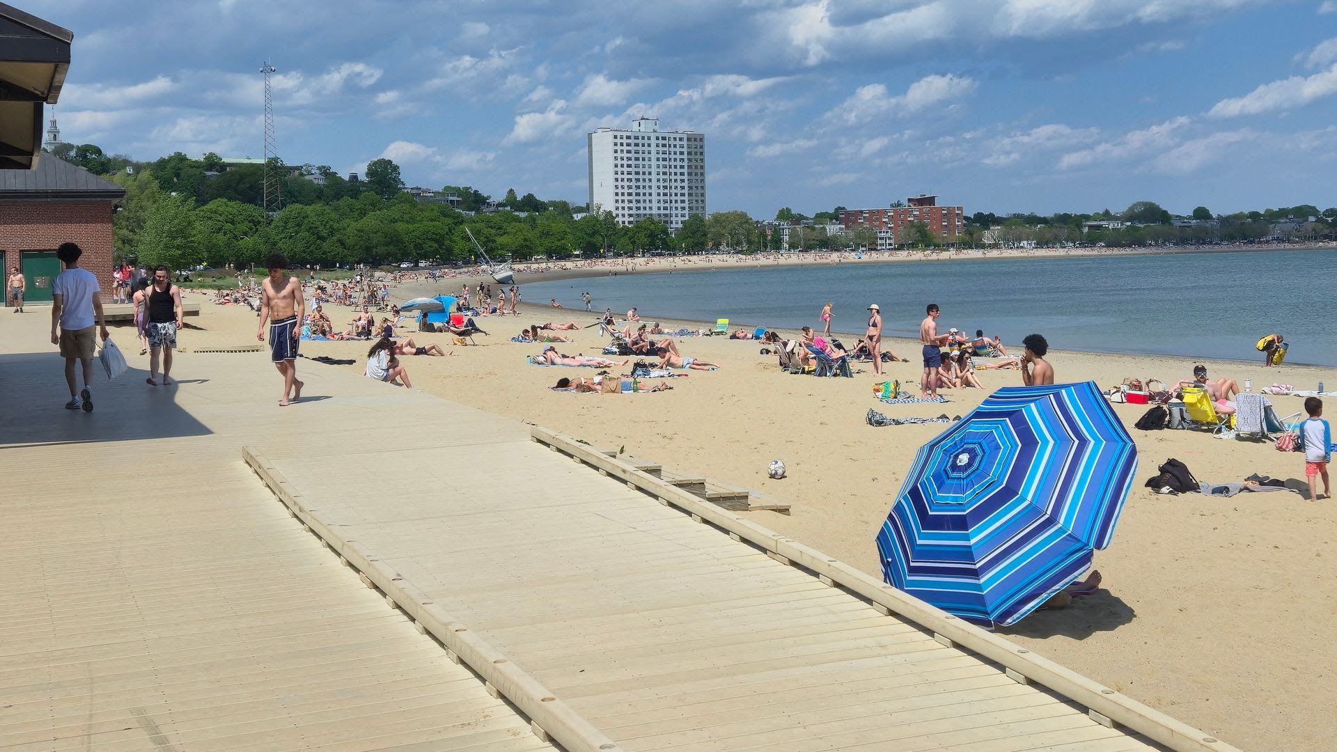 Busy beach under a blue sky: a wooden boardwalk on the left, crowds sunbathing and swimming, a blue striped umbrella in the foreground, and calm water with a white high-rise in the distance.