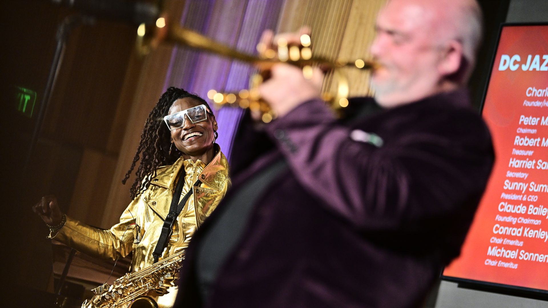 Smiling female saxophonist in gold outfit and clear glasses watches a male trumpet player in purple jacket performing at a D.C. Jazz event with red program board in background.