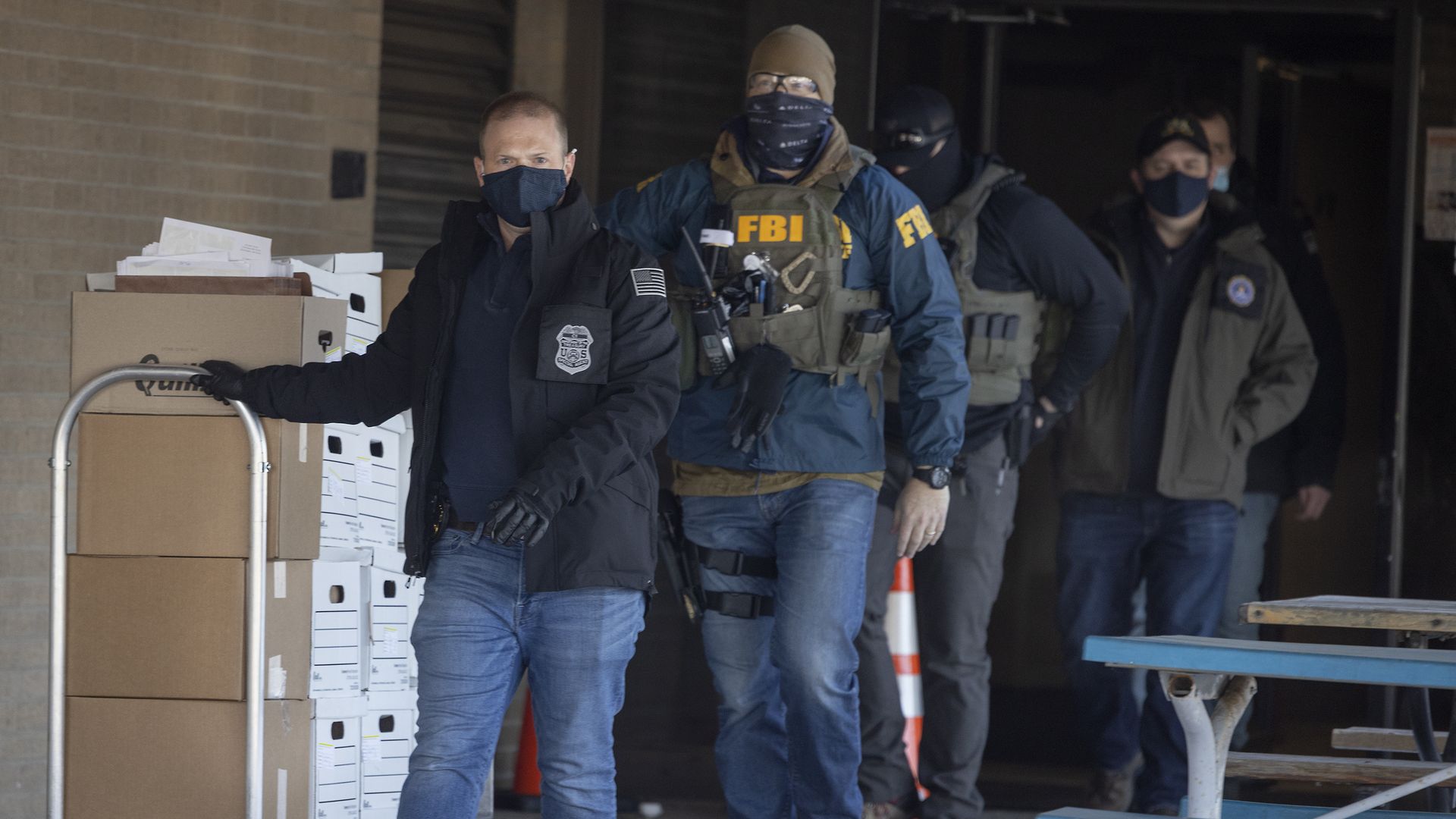 FBI agents wearing jackets and face masks, one pushing a cart loaded with boxes and documents outside a building with snow on the ground and a blue picnic table nearby.