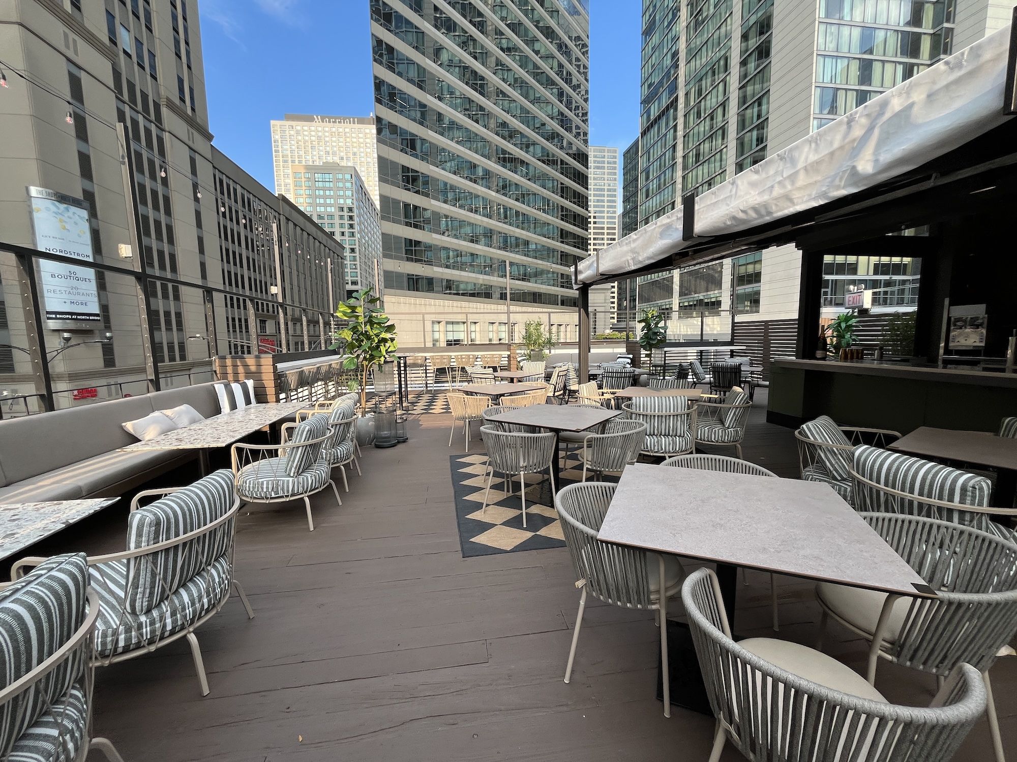 Urban rooftop cafe or terrace with many round tables and striped wicker chairs, string lights, plants, and modern glass towers surrounding under a clear blue sky.