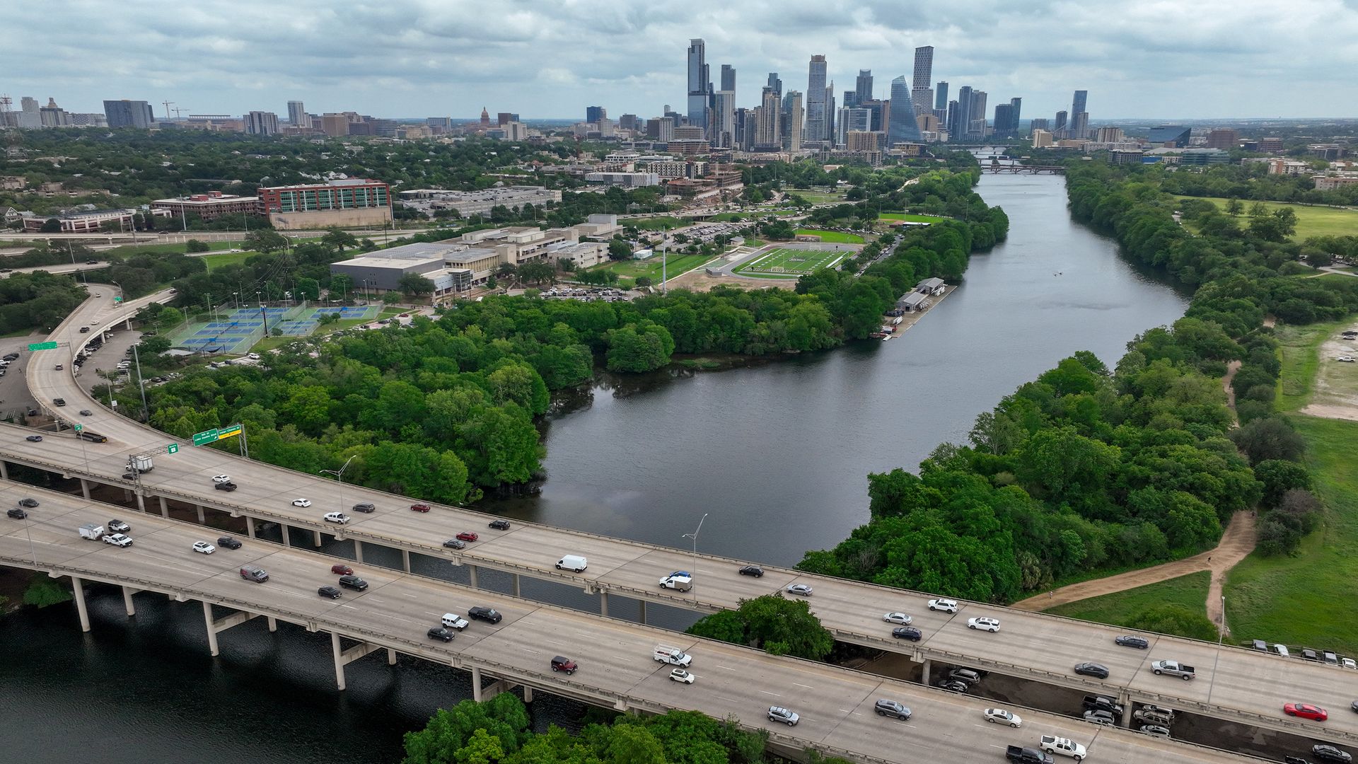 Aerial view of a city with a winding river lined by green parks, a dense downtown skyline of tall glass buildings, and a broad multi-lane highway in the foreground.