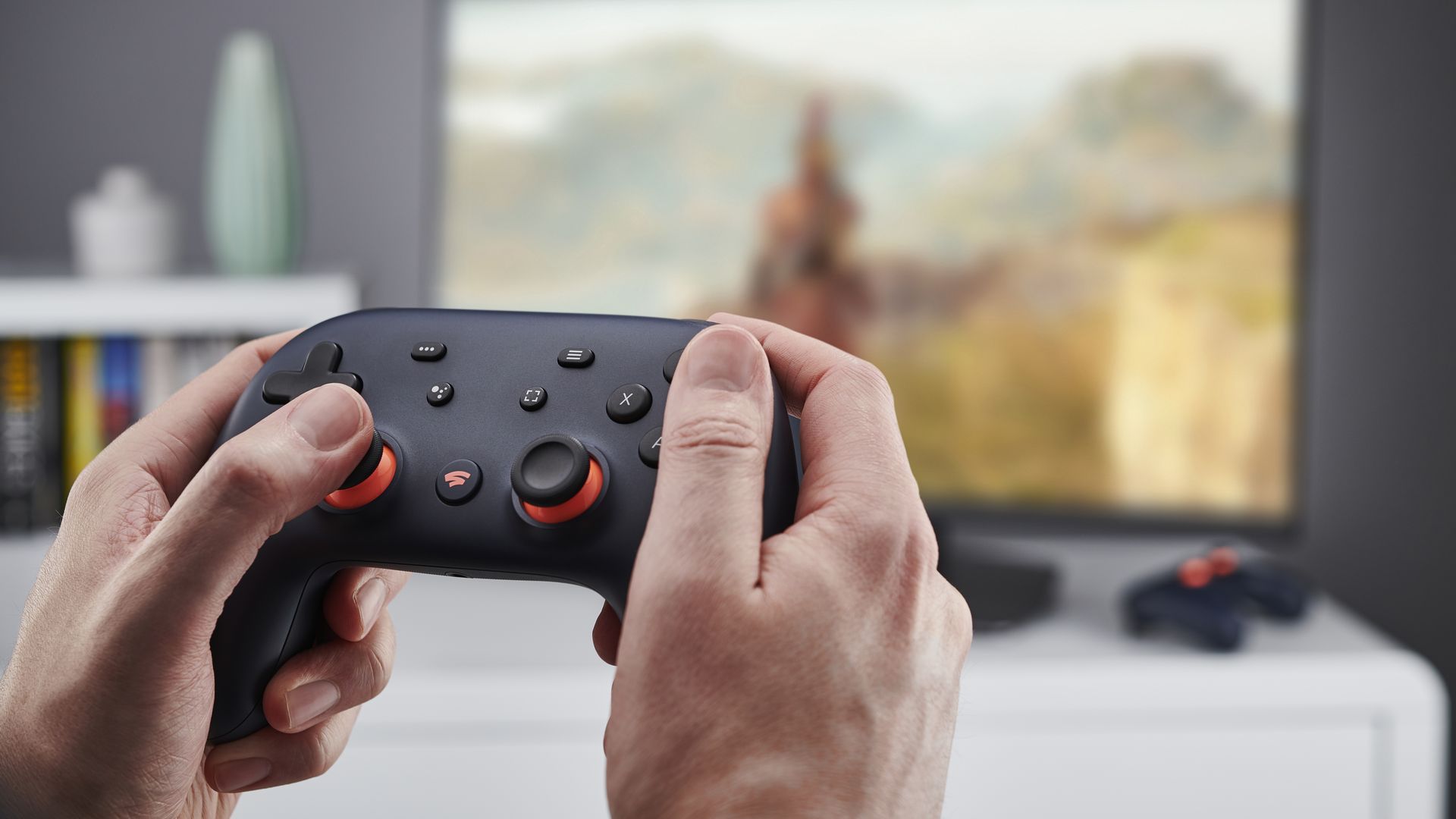 Photo of  a person's hands as they are holding a black game controller. A blurred-out TV can be seen in the background