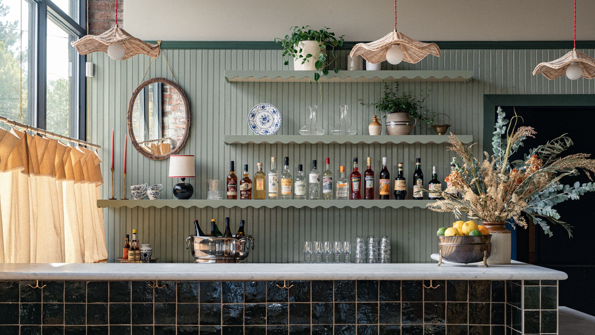 Cozy bar with green paneled walls, wooden stools, shelves of liquor bottles and glassware, hanging wicker lamps, potted plants, a fruit bowl, and dried floral arrangement on a tiled counter.