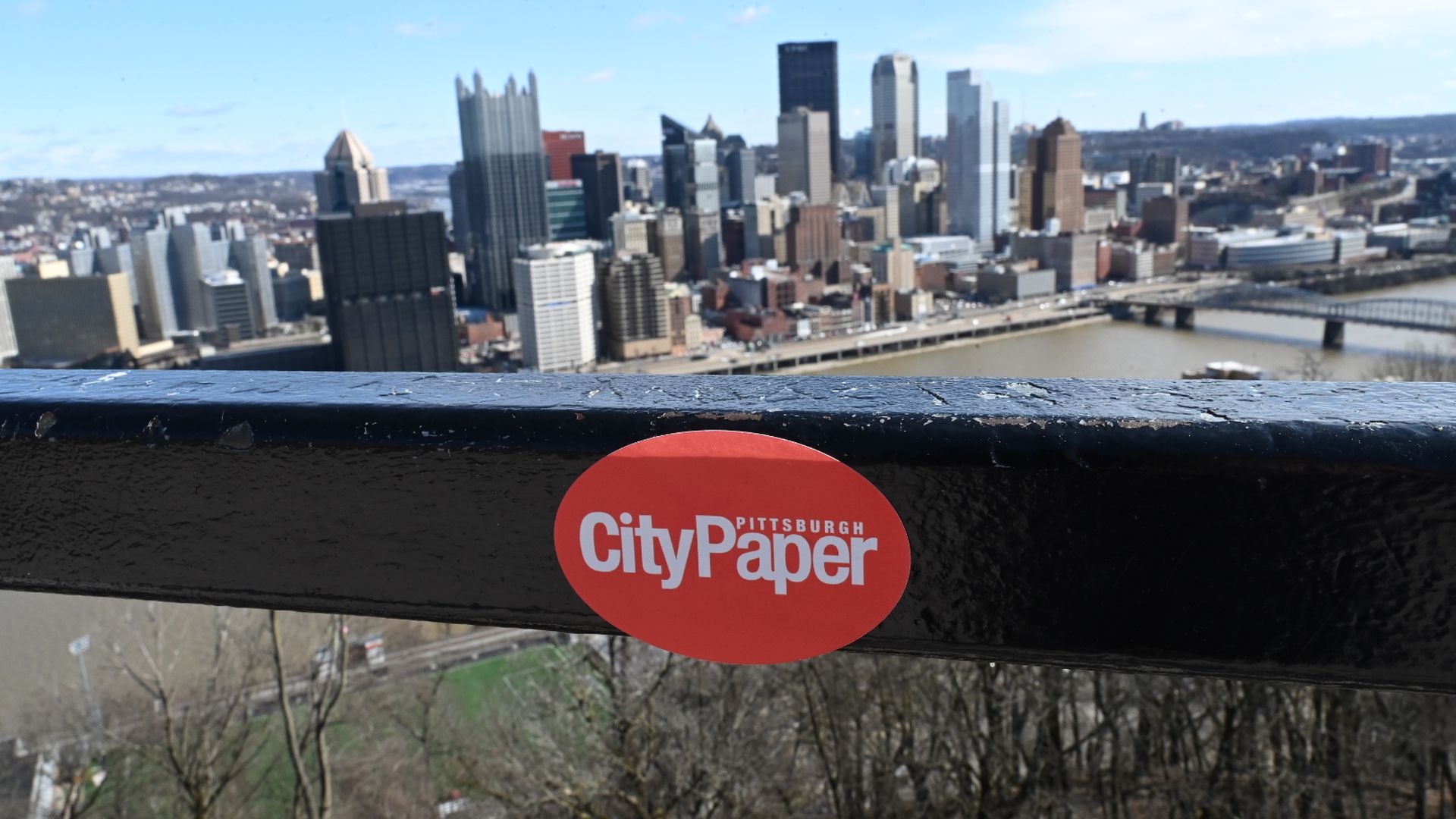 Pittsburgh skyline with a river and bridges under a blue sky. In the foreground, a black railing with a red oval sticker reading "CityPaper".