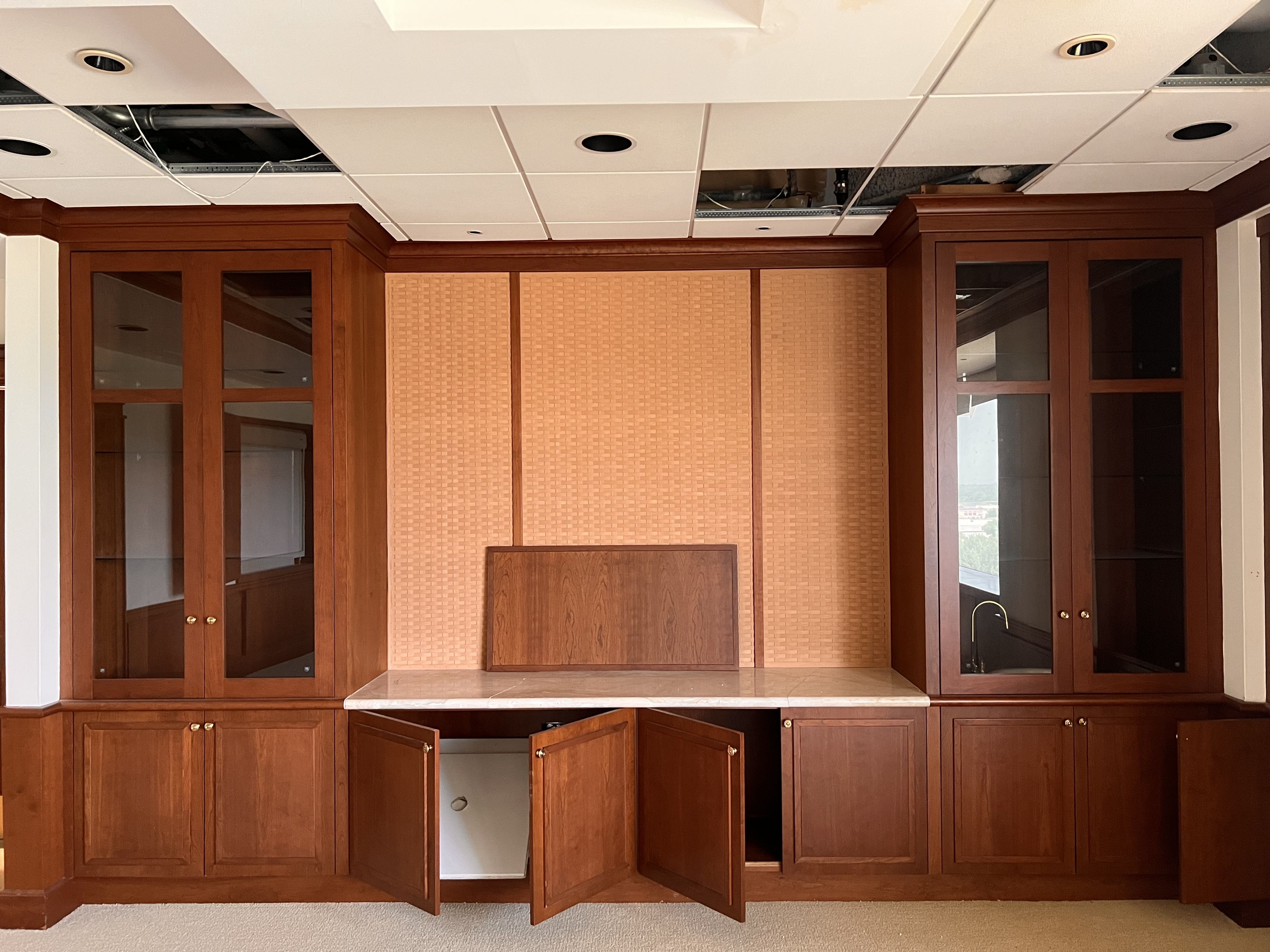 Dark wood cabinetry with glass doors, a beige woven center panel, and a marble countertop. Open ceiling tiles above reveal wires and exposed ducts.
