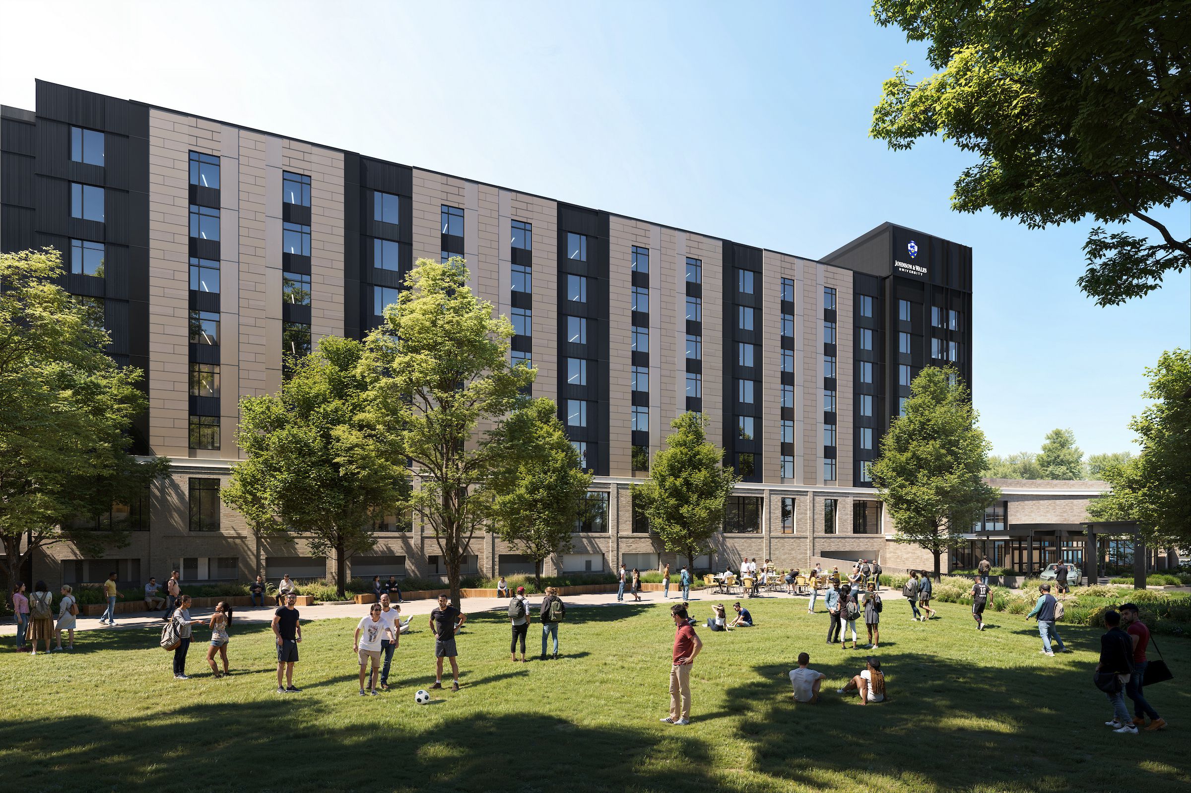 Modern university building with dark panels and tan accents behind a green lawn. People walk, talk, and sit on the grass; a Johnson & Wales University crest is visible on the right.