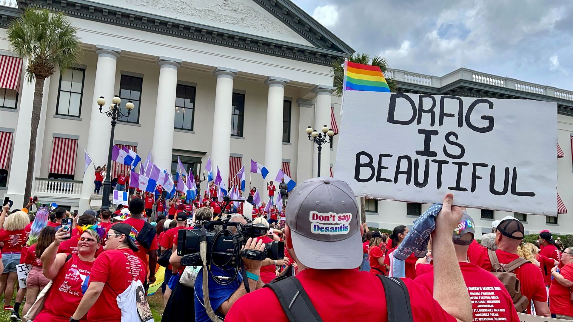 A person wearing a hat that says "Don't say DeSantis" stands in front of a protest at the Florida State Capitol in Tallahassee, holding up a sign that says "Drag Is Beautiful."