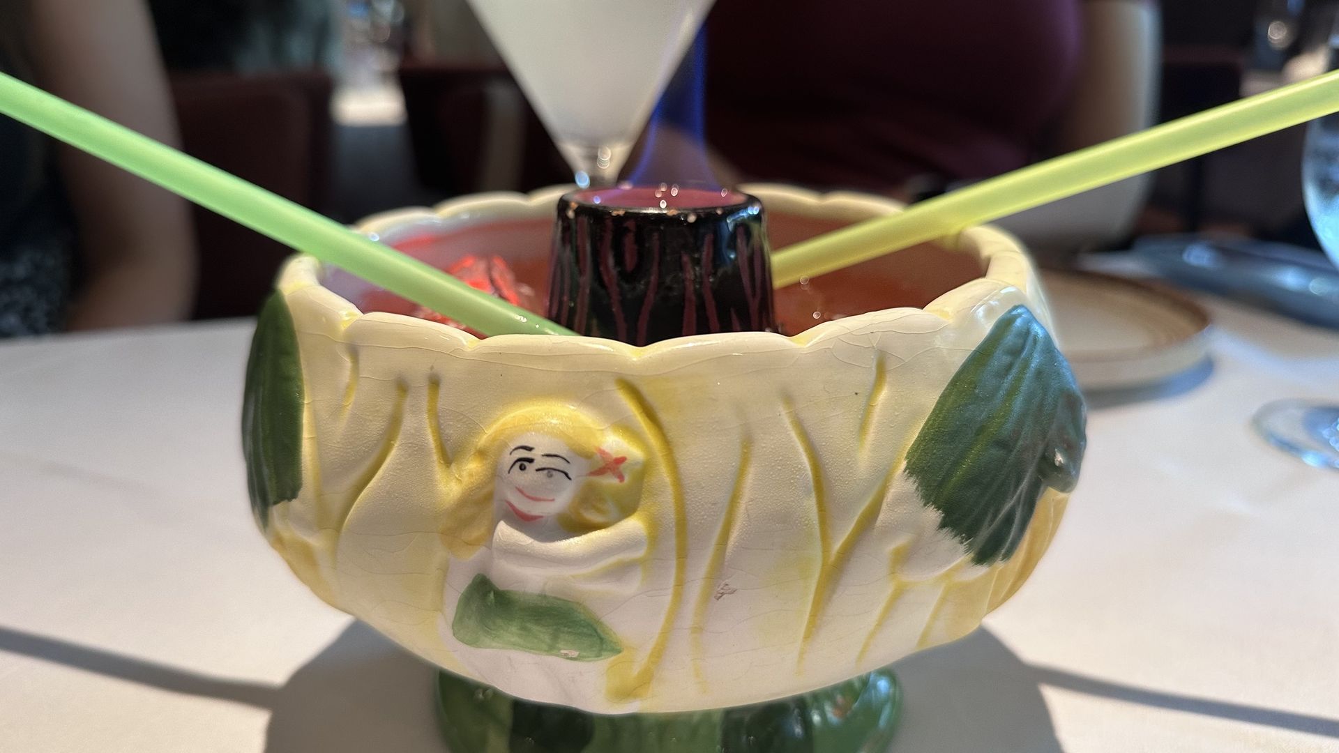 Ceramic bowl with yellow and green hula dancer decorations, holding a flaming drink with two yellow straws on a white tablecloth in a restaurant setting.