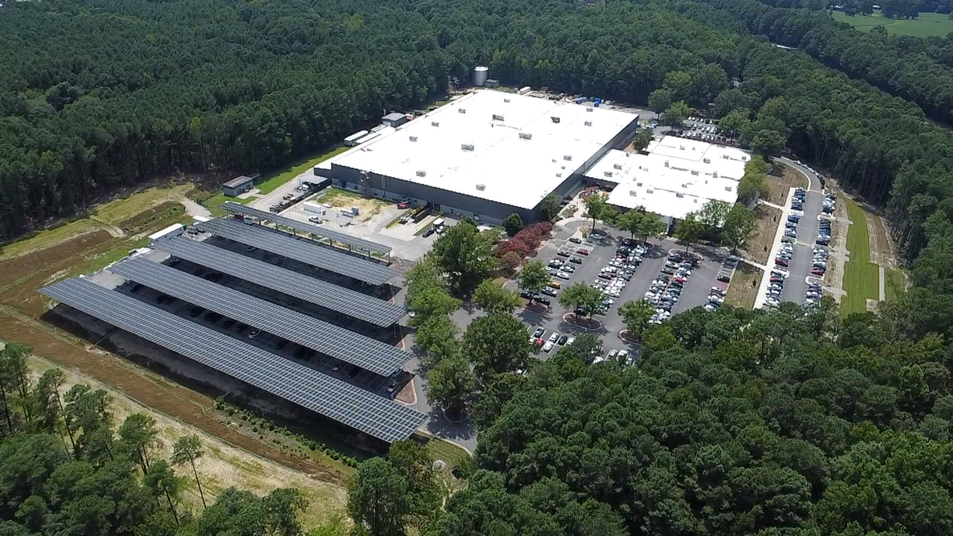 Aerial view of an industrial complex in a forest: three large white-roof buildings, a long solar-panel carport, and a spacious parking lot with many cars, all bordered by dense trees.