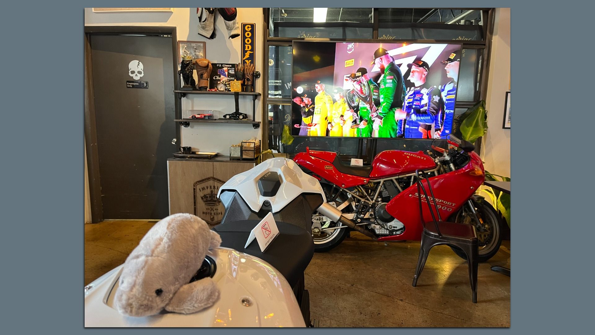 Indoor motorcycle showroom featuring a red Ducati sport bike beside a white futuristic bike, a gray door with a skull sticker, shelving with gear, and a large screen displaying color-clad racers.