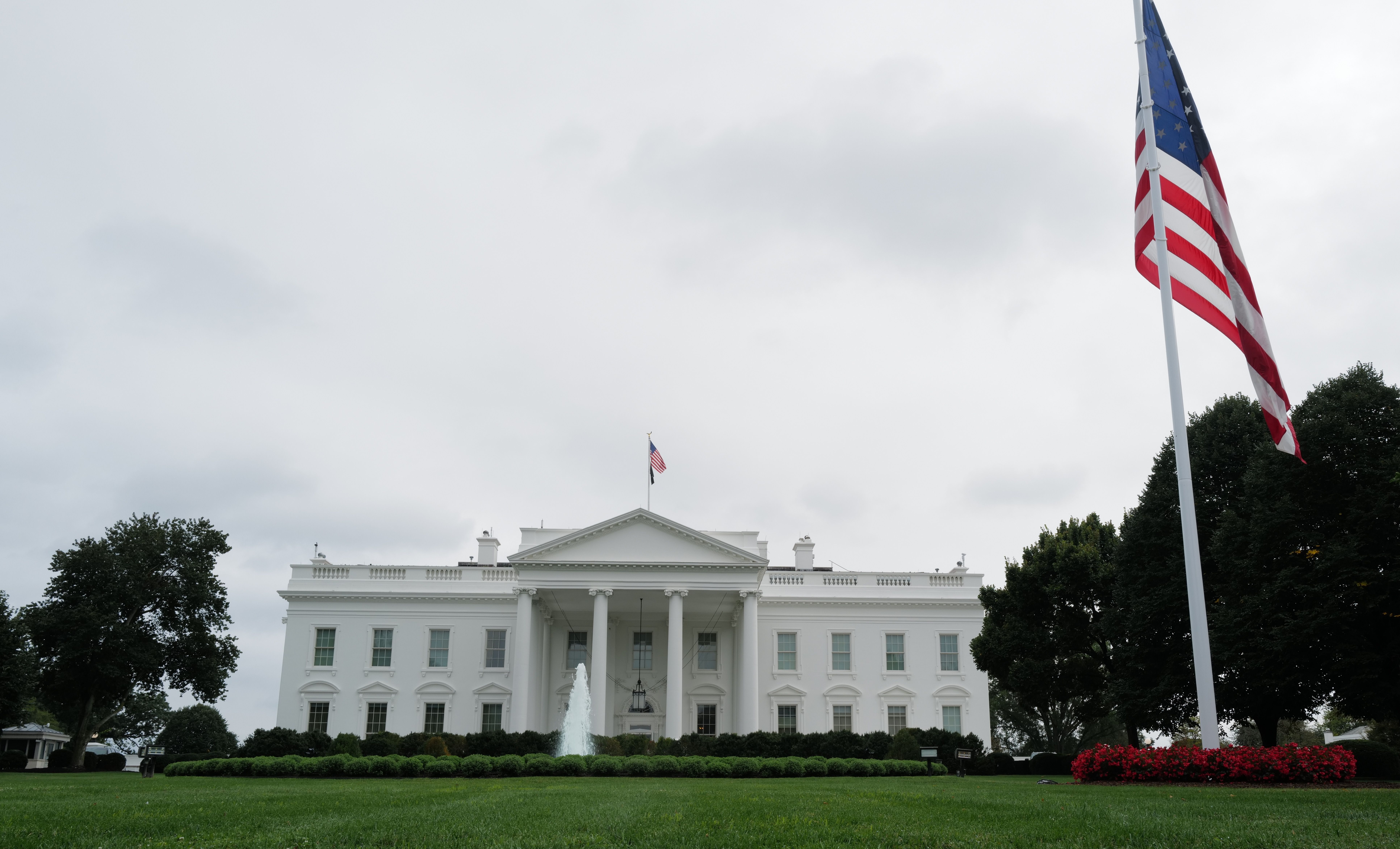 The White House with an American flag flying on a pole in the foreground, green lawn, bushes, and trees surrounding it under a cloudy sky.