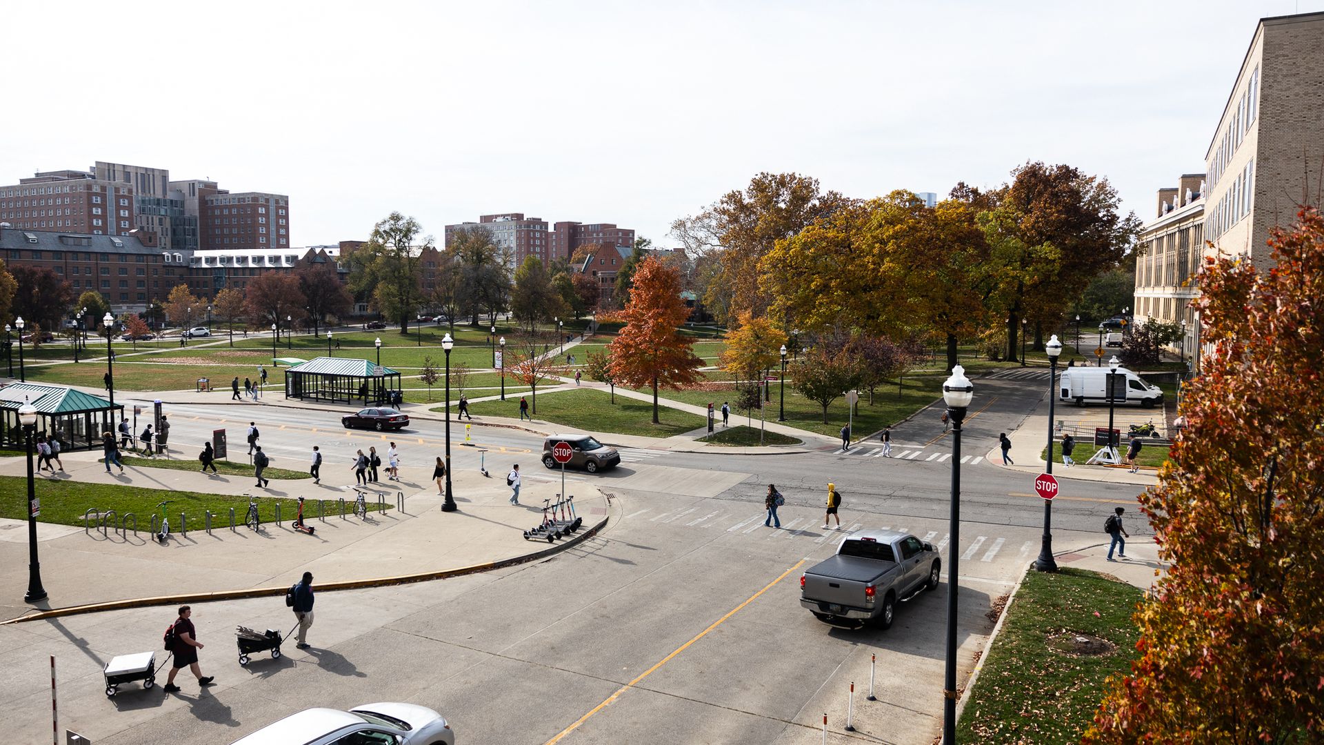 Panoramic campus view: brick buildings left, a large beige brick building right, vibrant fall trees, green lawns, and pedestrians crossing streets on a sunny day.