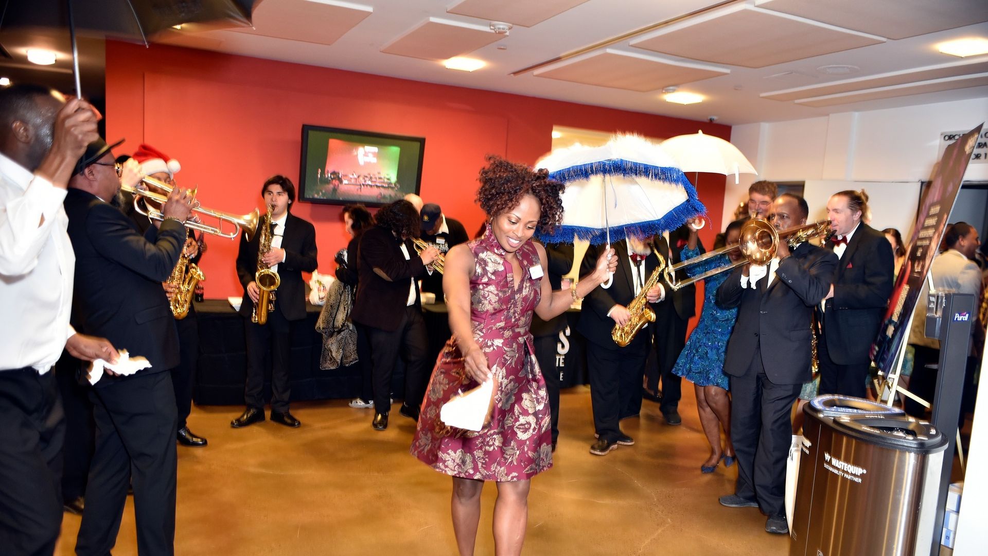 Woman in red floral dress dancing with a white and blue umbrella among musicians playing brass instruments in a lively indoor setting with red walls and a TV screen.