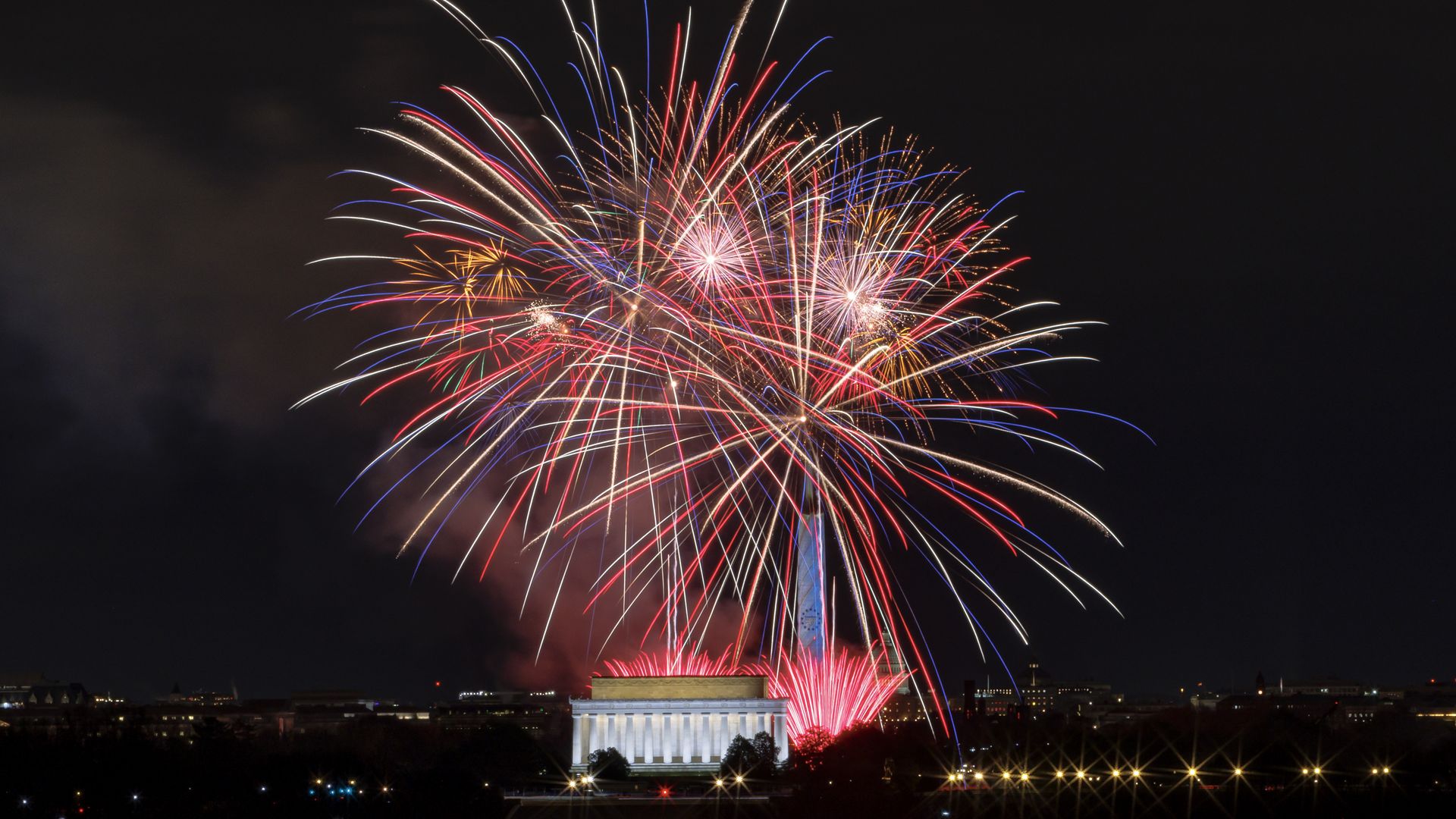 Night scene of fireworks in red, white, and blue bursting over the National Mall, with the Washington Monument behind a lit Lincoln Memorial and city lights along the horizon.