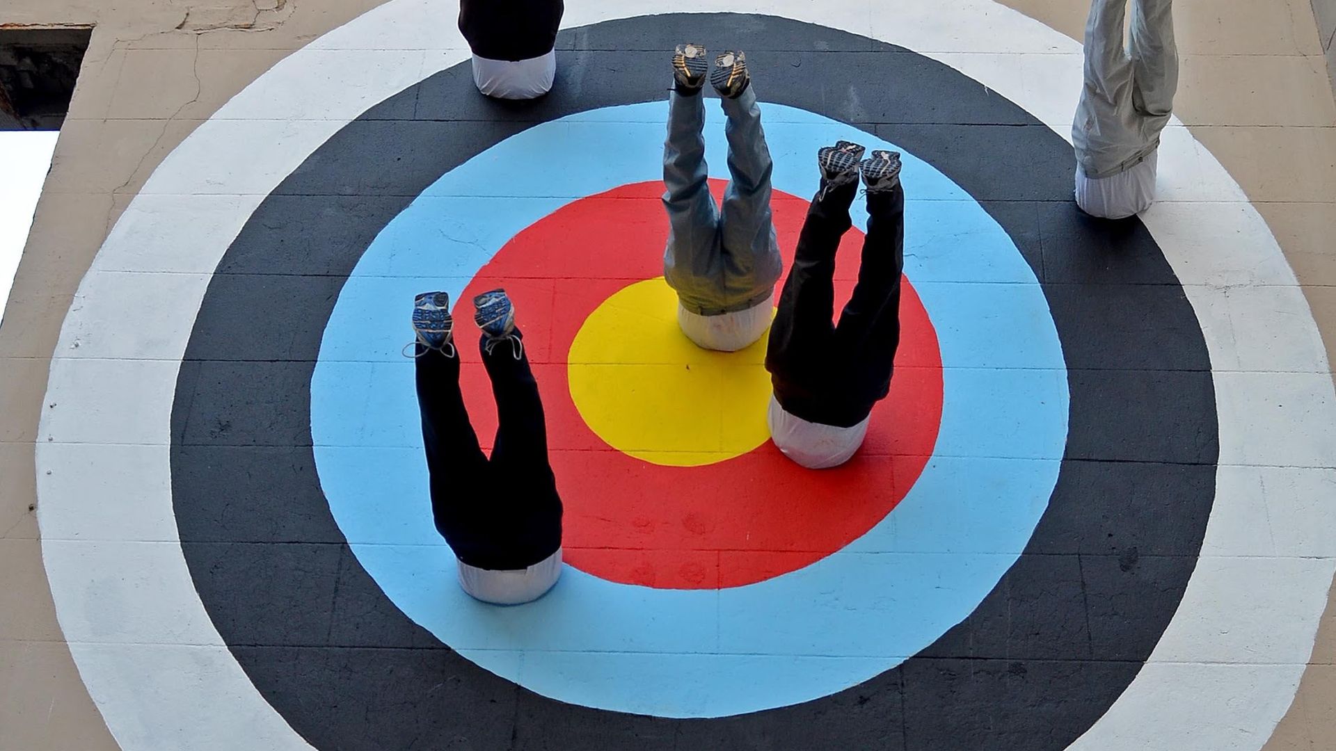 Overhead view of a painted archery target on a concrete floor; several people lie on their backs on the rings with legs raised, wearing sneakers.