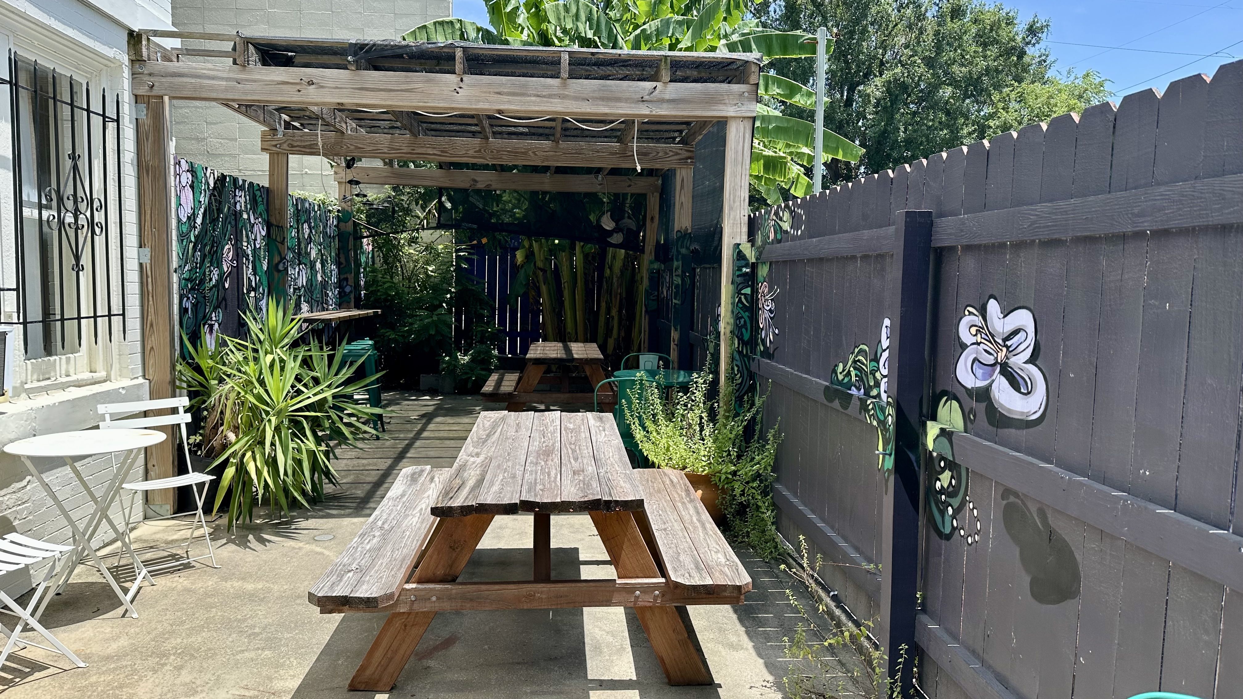 Backyard alley with a weathered wooden pergola over a long picnic table. Graffiti murals on dark fences, lush plants along the sides, and a small white metal bistro set. Clear blue sky above.